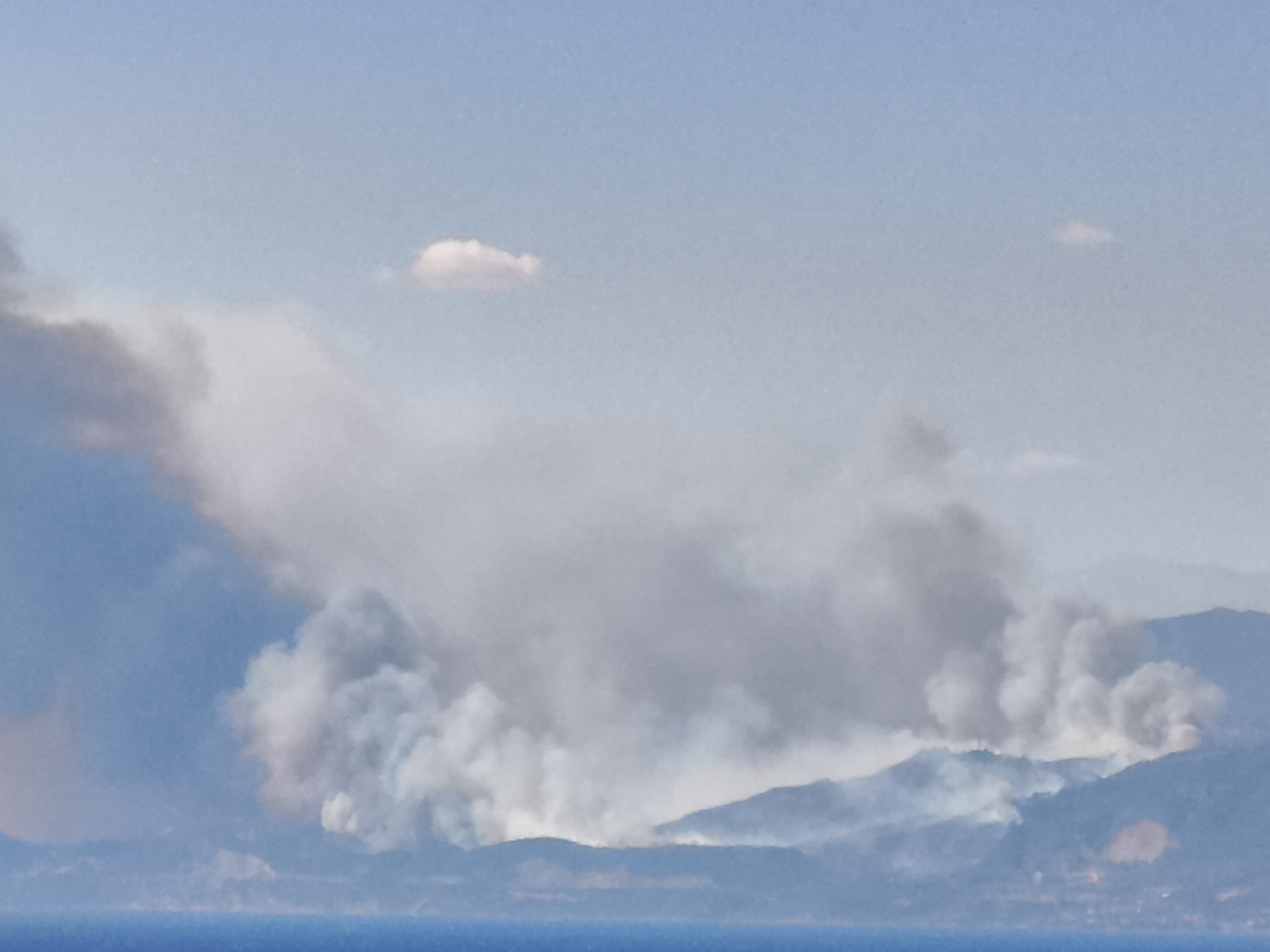 Wildfire smoke rises over Aegio's coastline, visible from the waterfront