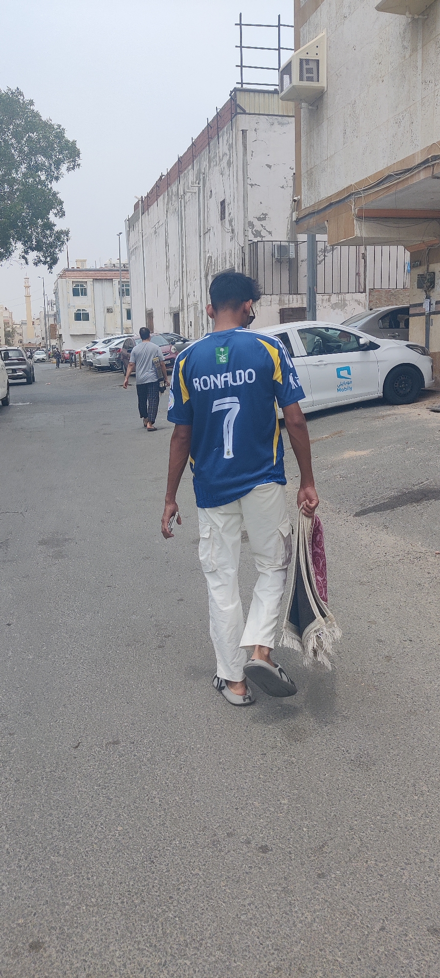Pedestrians walk Jeddah street, man carries prayer rug