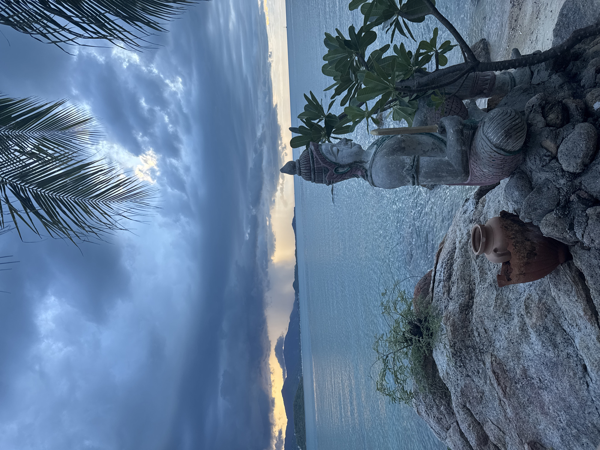 Ko Samui at dusk: coastal statue, stormy sunset, warm lamp indoors