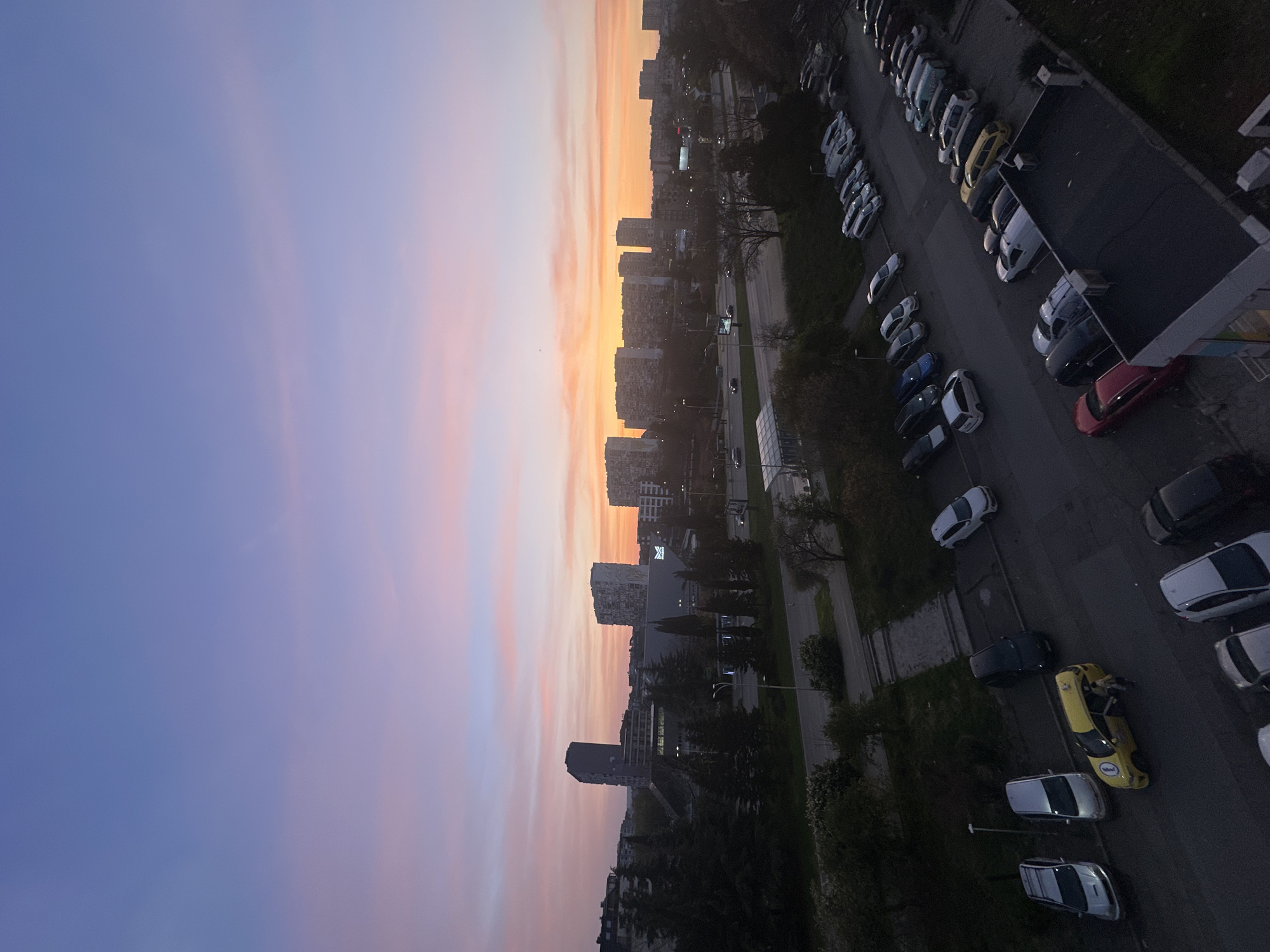 Urban dusk scene shows apartment buildings, parked cars, greenery