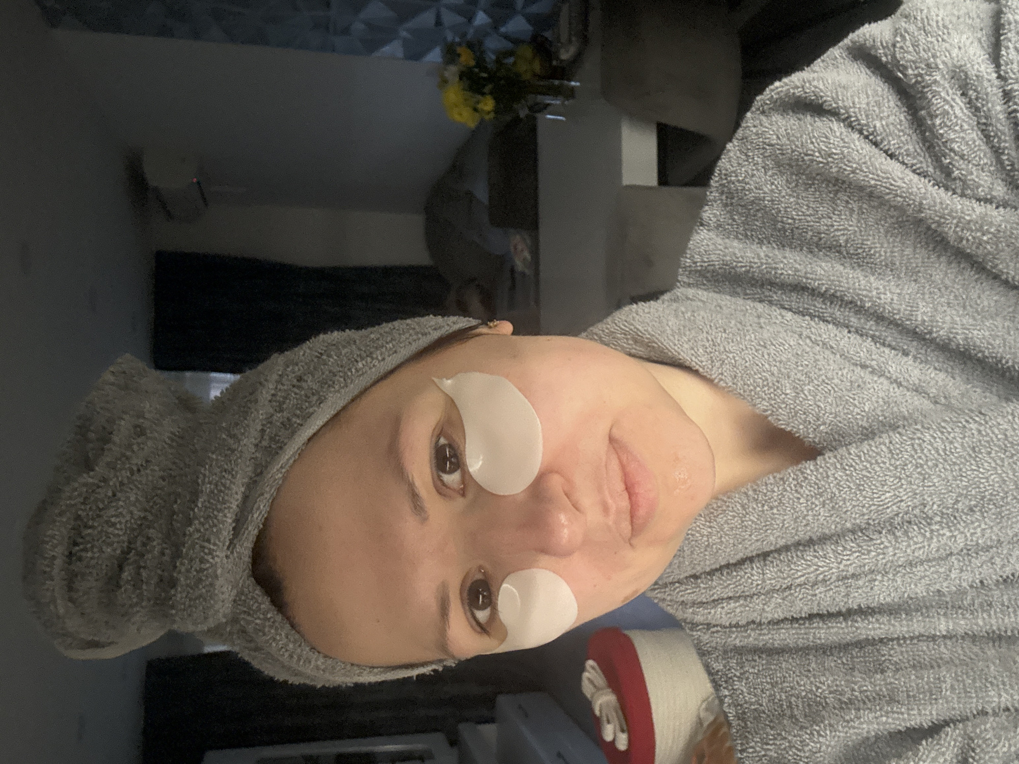 Woman in bathrobe with facial patches holds mug indoors