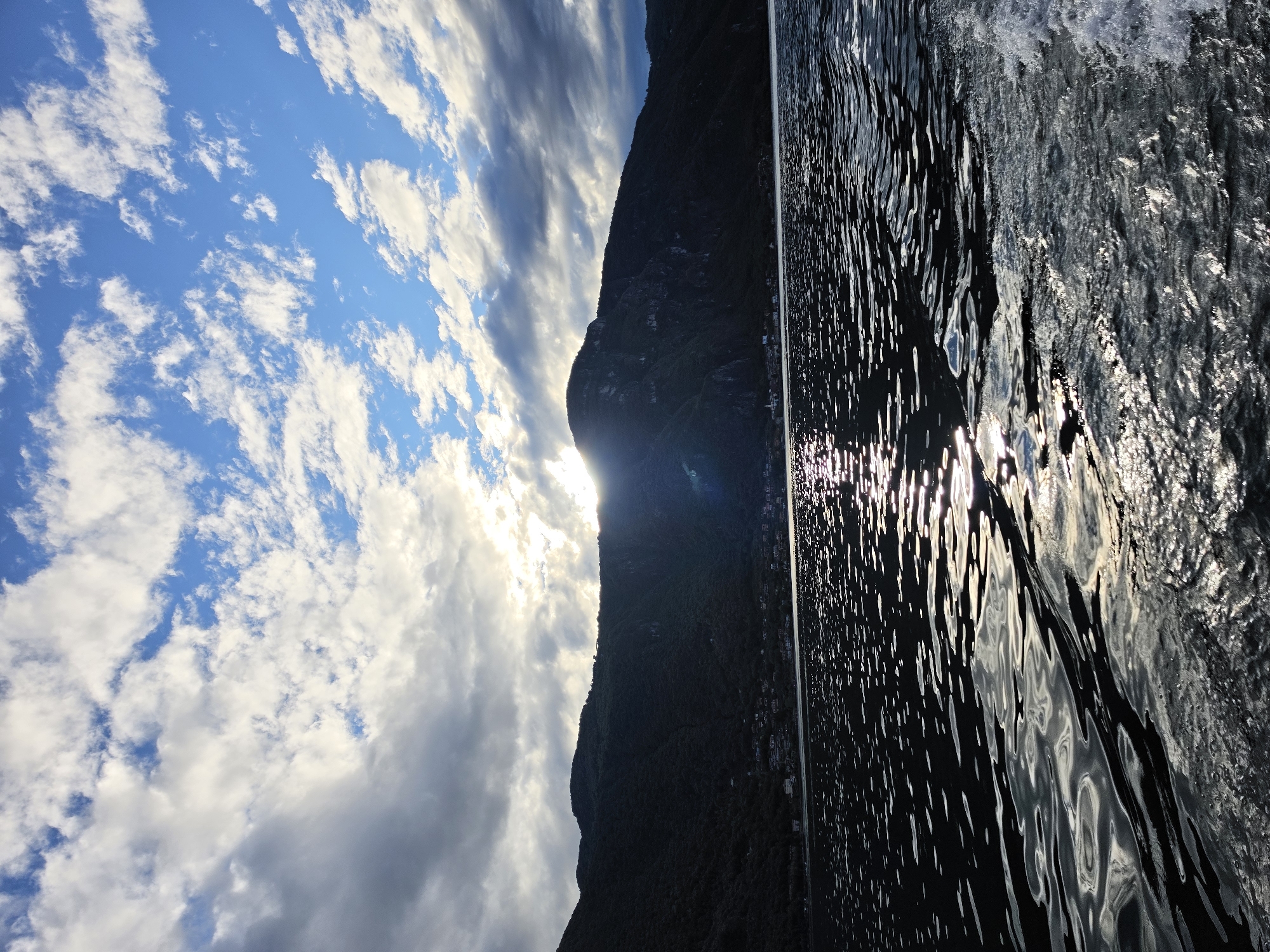 Lake Como shoreline from the water, sun cutting through afternoon clouds