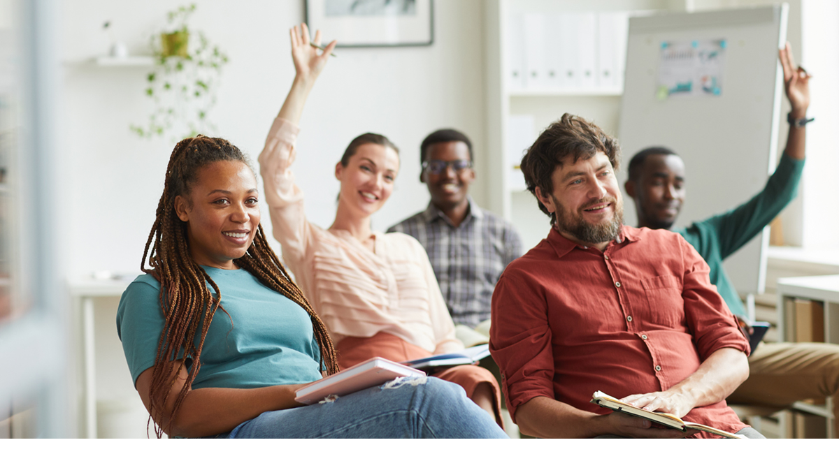 A cohort of coworkers sits in a classroom-like environment. Several employees are contributing to the conversation by raising their hands, while others take notes.