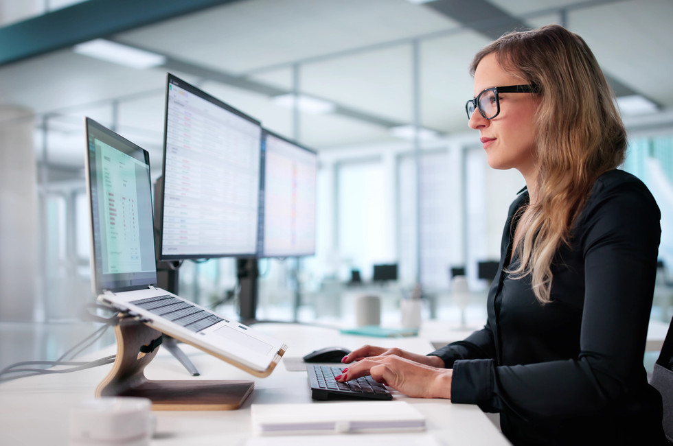 Financial controller at her desk in a bright office, reviewing accounts payable data on two monitors and a laptop