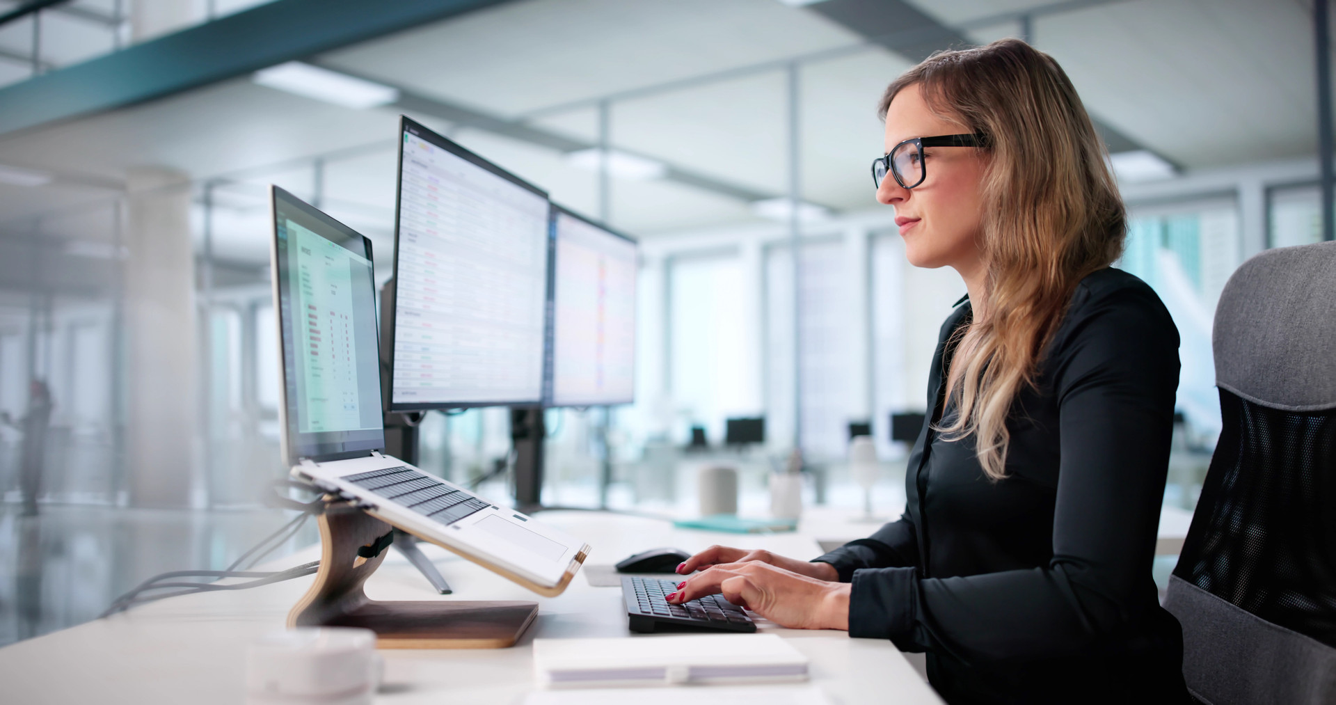 Financial controller at her desk in a bright office, reviewing accounts payable data on two monitors and a laptop