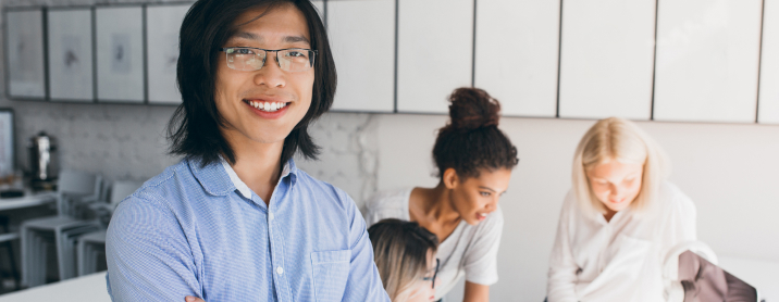 Young smiling engineer making eye contact with the camera while his team works behind him. 