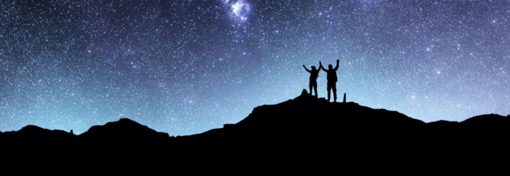 Two people standing on a mountain top in the distance looking at a beautiful starry night