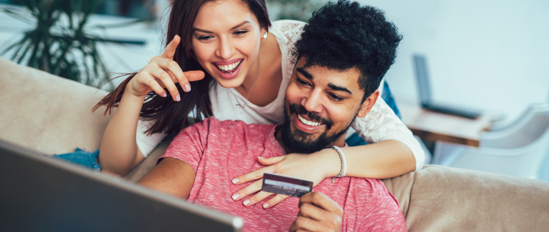 Happy young man sitting on a couch shopping on his laptop with his wife behind him leaning over his shoulder