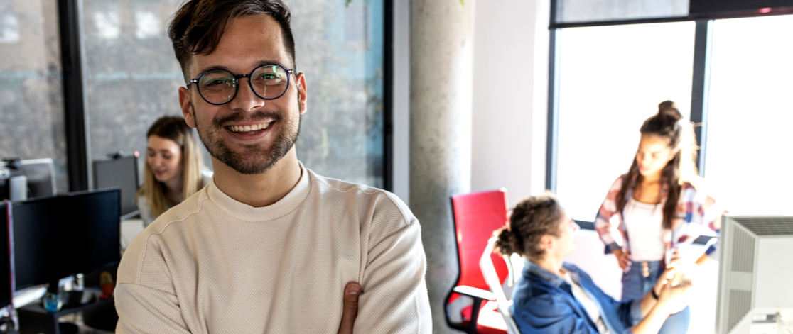 Smiling young man standing in office with his coworkers sitting behind him at their desks