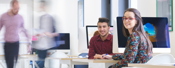 Two programmers sitting in a busy office and smiling at the camera