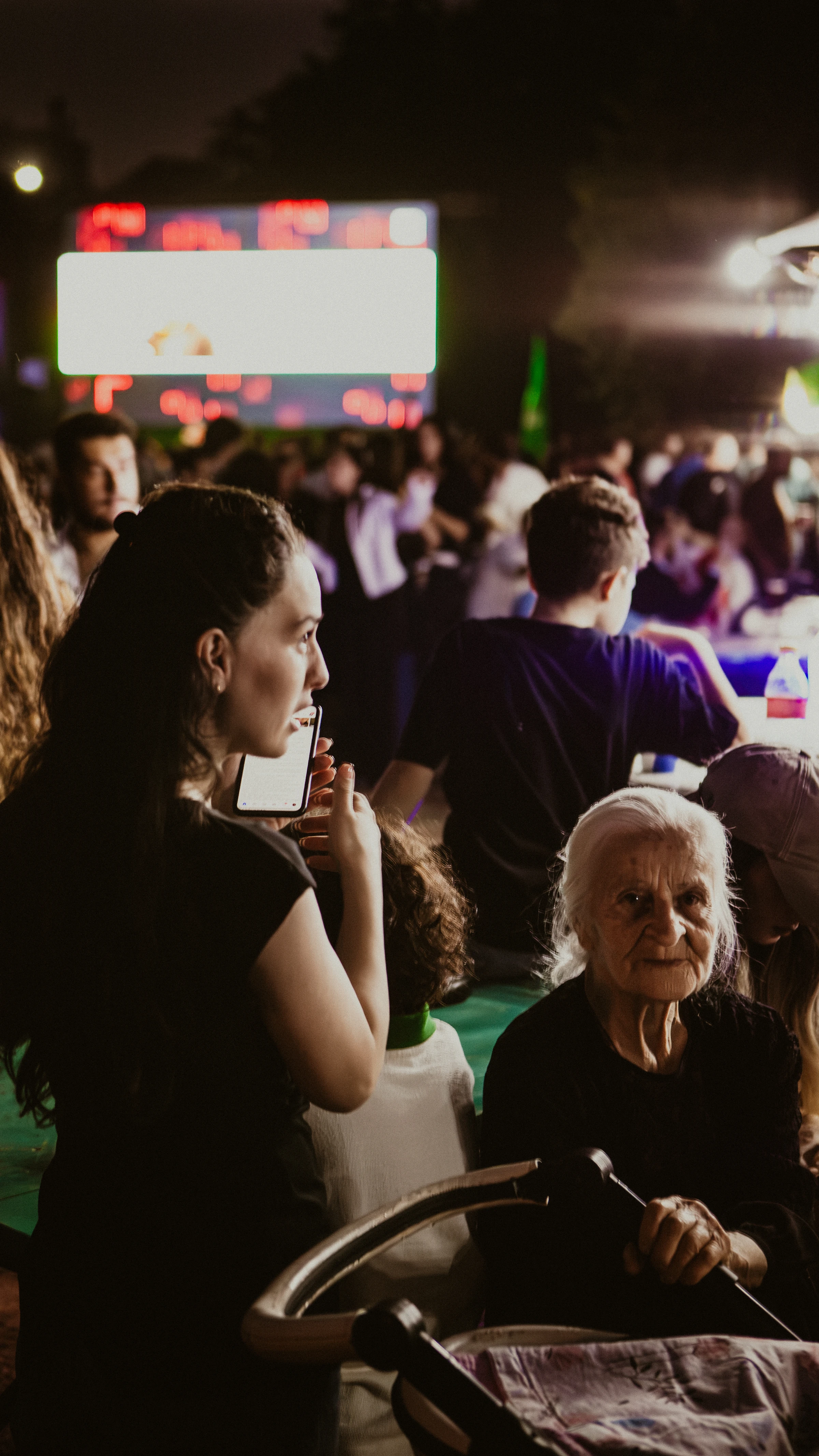 Woman holding a smartphone in front of her face at a nighttime outdoor event with a large display screen visible behind the crowd.