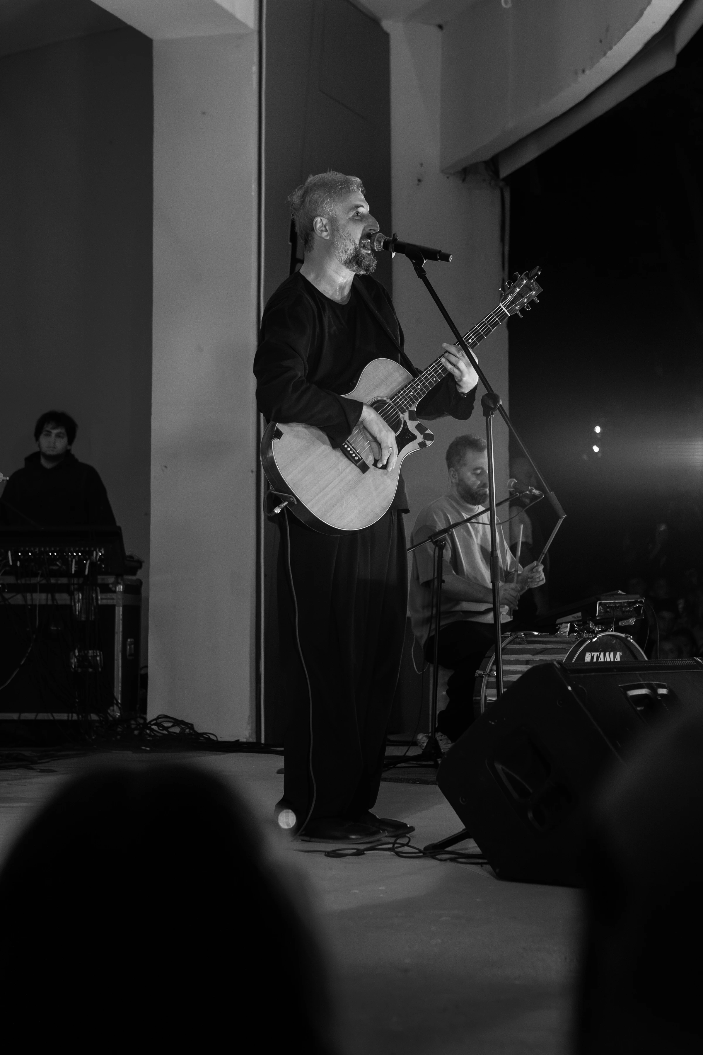 A man sings into a microphone while playing an acoustic guitar, with a keyboardist and drummer visible behind him on stage.