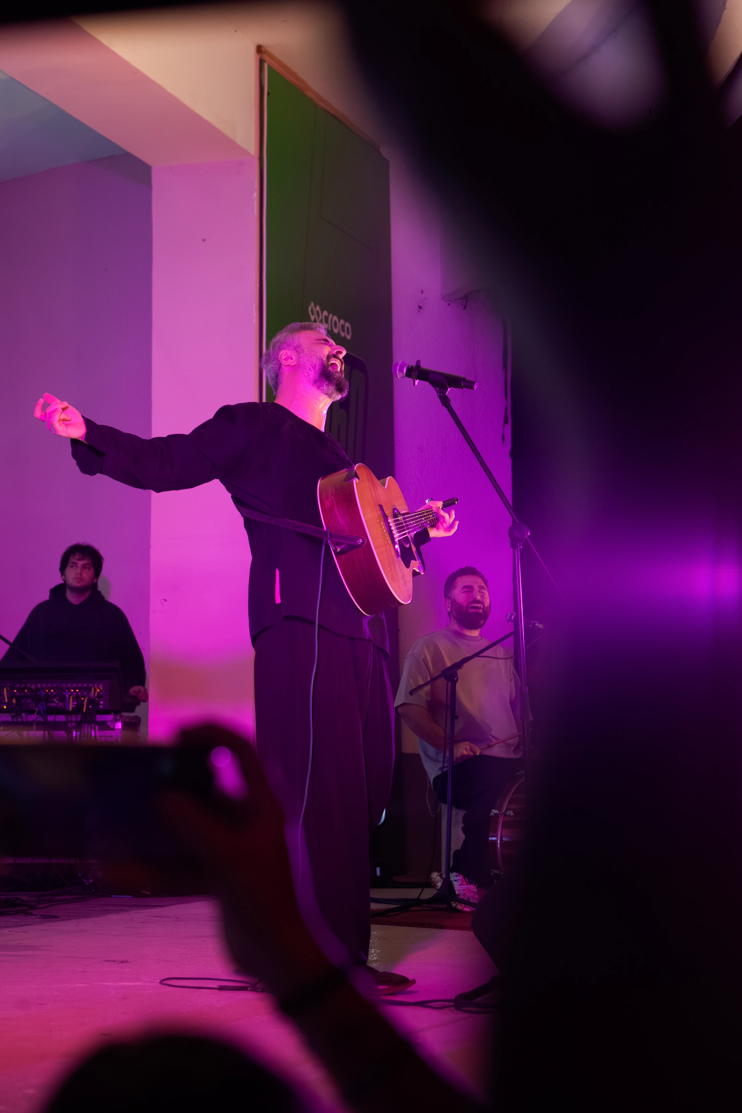 A performer with an acoustic guitar stands on stage with raised arm under purple lighting, with musicians visible behind him.