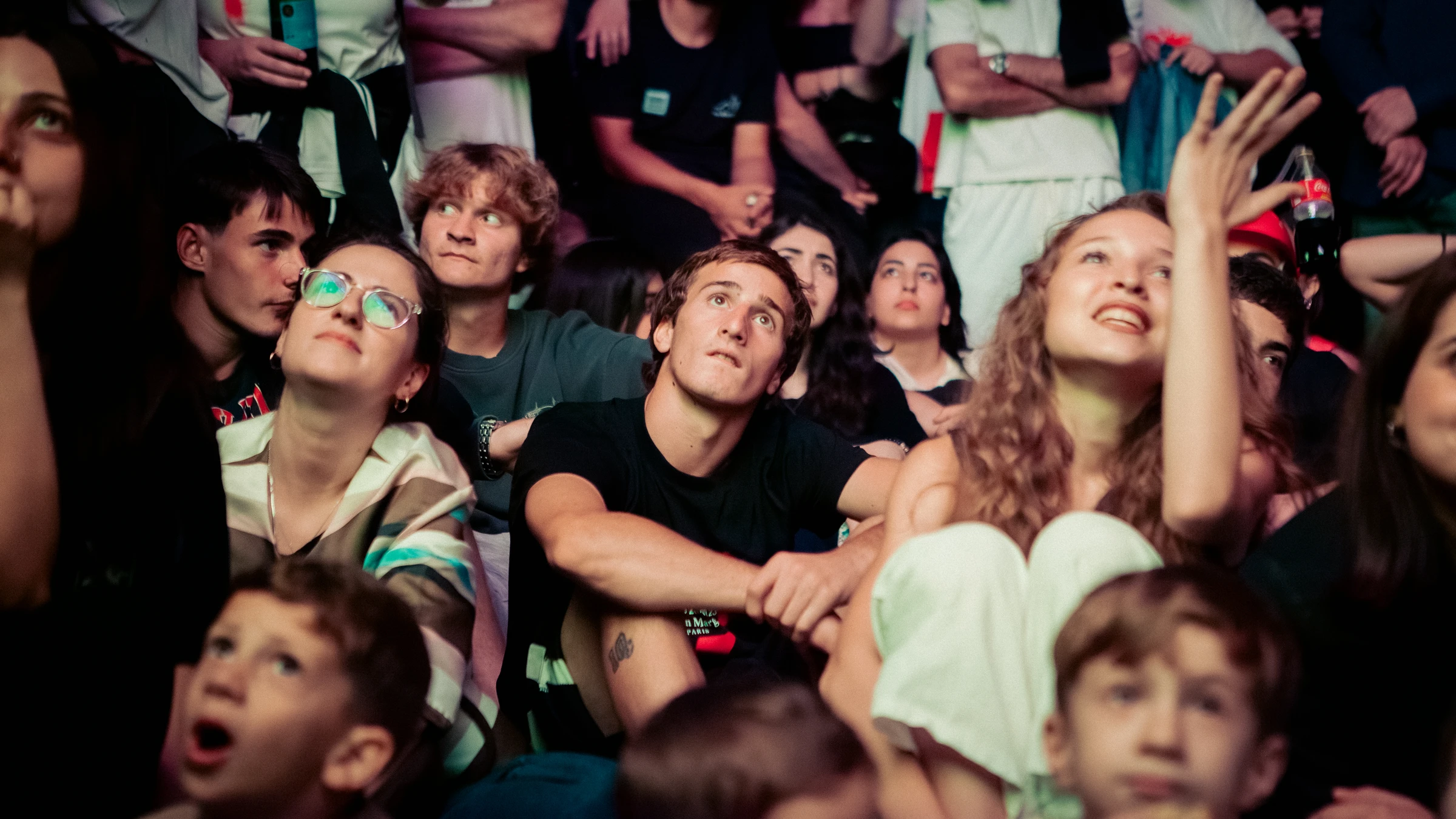 A crowd of young spectators at a night concert gazes upward with expressions of awe and concentration.