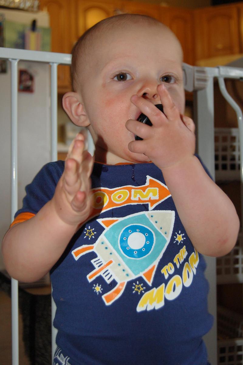One-year-old Calvin, dressed in spaceship themed pajamas, looks at the camera while trying to shove some unidentified round shape into his mouth. The pajamas are dark blue with orange trim around the sleeves and neck and a stylized spaceship on the front.