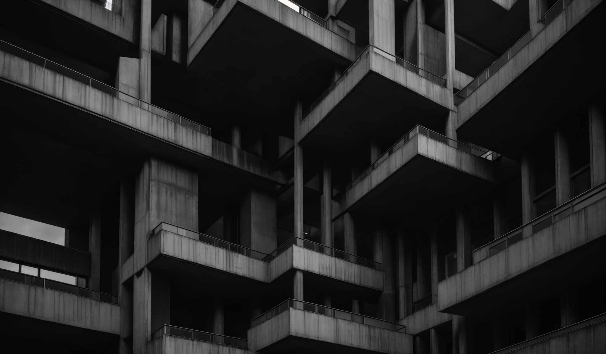 The image showcases a black and white architectural photograph of a building. The building features multiple interconnected balconies and platforms, creating a maze-like structure. The architectural style is reminiscent of Brutalism, characterized by its raw concrete construction. The image emphasizes the geometric shapes, the play of light and shadow on the surfaces, and the intricate interplay of the balcony levels.