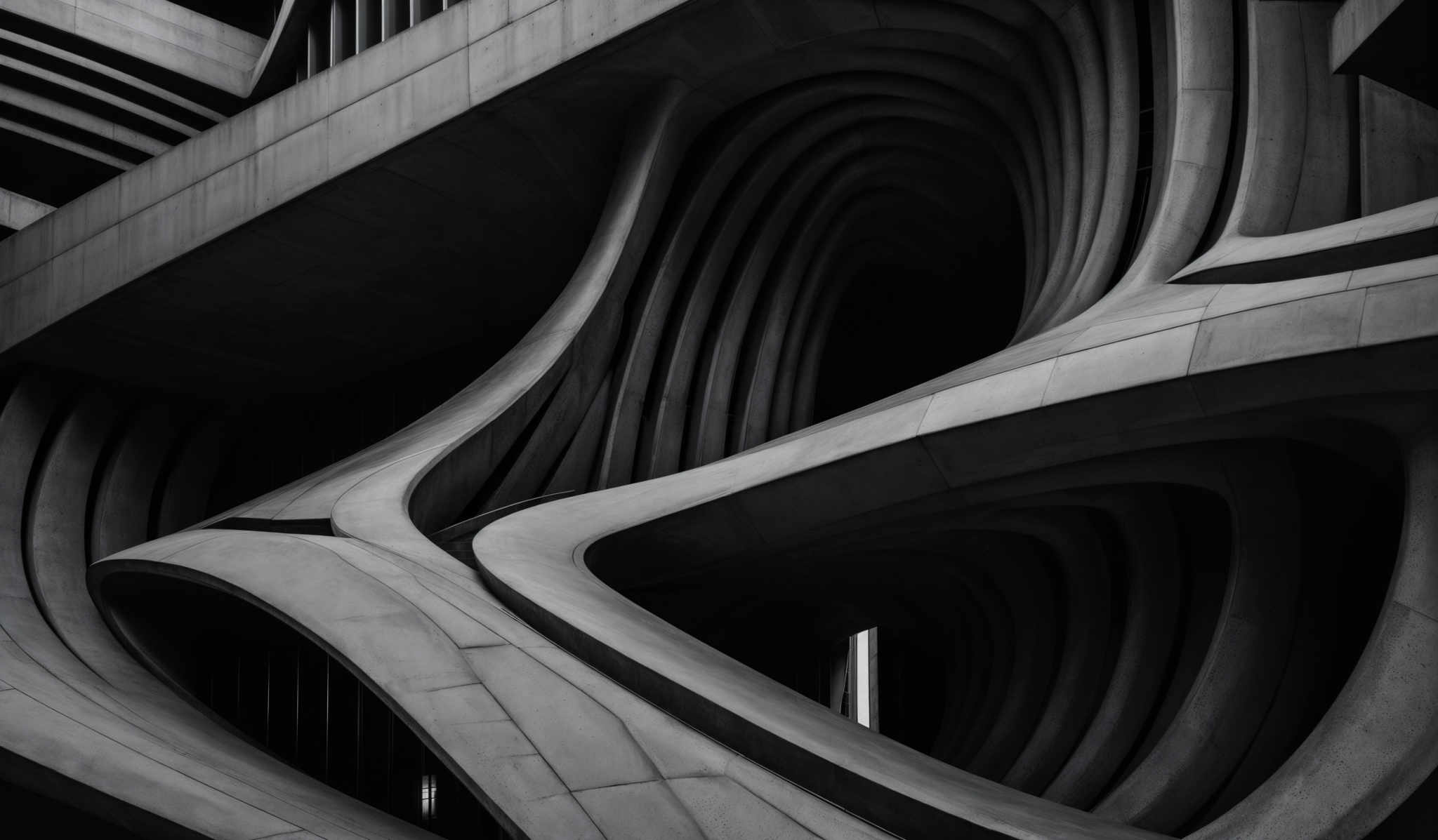 The image showcases a black and white architectural photograph. It features a building with intricate curved and wavy concrete structures. The design is reminiscent of flowing water or organic forms, with multiple arches and overlapping layers creating a sense of depth and movement. The play of light and shadow accentuates the curves and adds a dramatic effect to the overall composition.