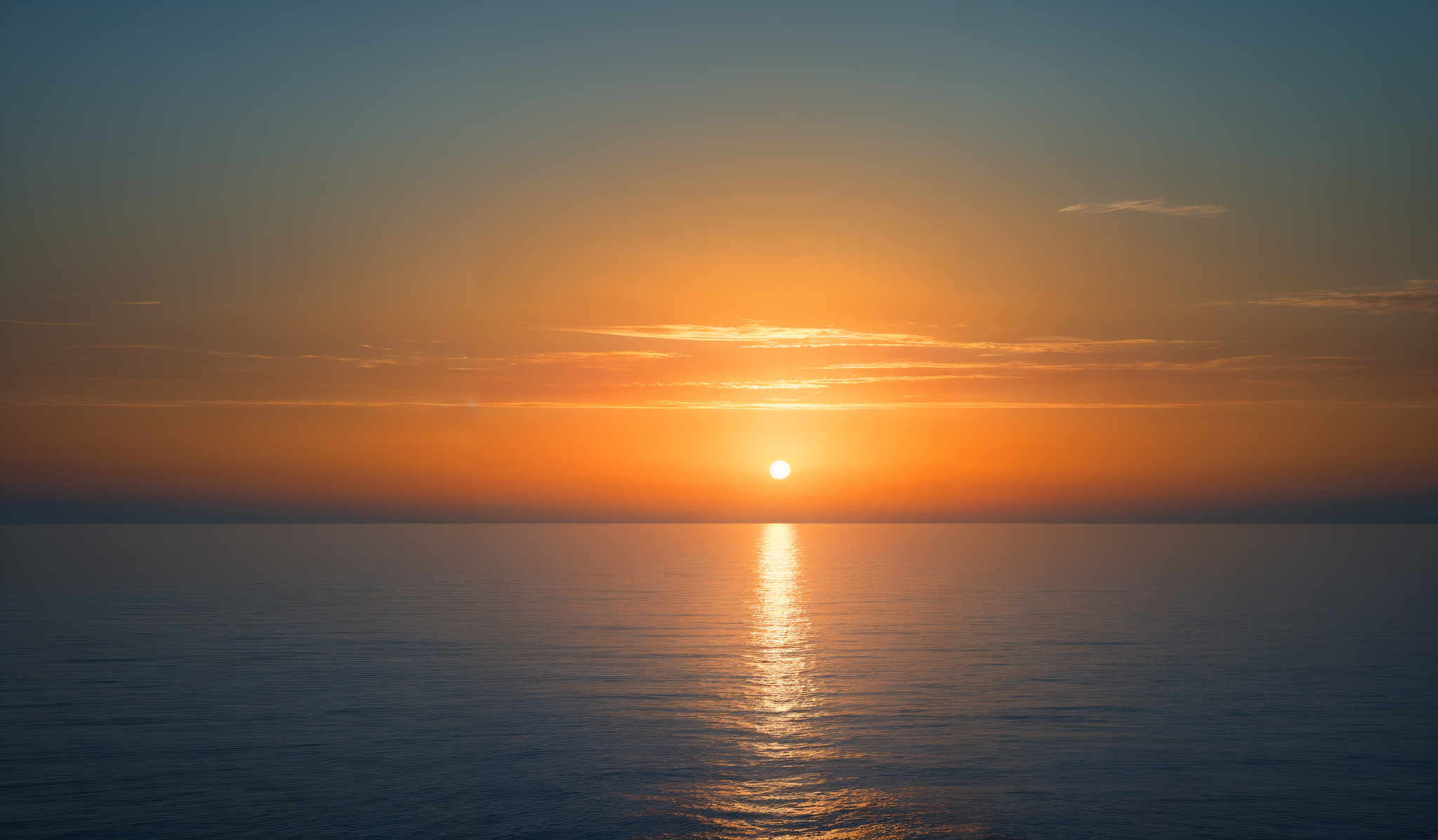 The image showcases a breathtaking sunset over the ocean. The sky is painted with hues of orange, yellow, and a hint of blue. The sun, positioned near the horizon, radiates a brilliant golden light that reflects on the calm waters below. The horizon line is clearly visible, separating the sky from the sea. The clouds, scattered across the sky, add depth and texture to the scene, with some appearing darker and others illuminated by the sun's glow.