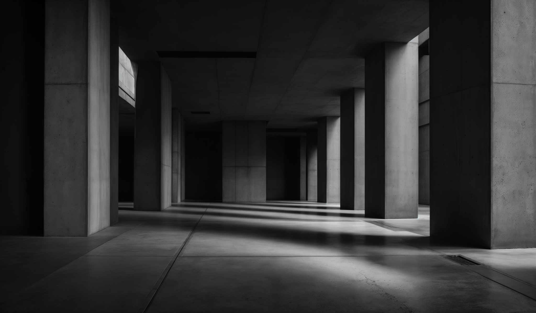 The image showcases a monochromatic, possibly black and white, architectural setting. It features tall, slender columns that extend from the floor to the ceiling. The columns are uniformly shaped and have a rough texture, possibly made of concrete. The floor is smooth, reflecting the columns and the light sources. The image captures a play of light and shadow, with the columns casting shadows on the floor. The perspective suggests a corridor or hallway, with columns on both sides, leading to a darker space in the distance.