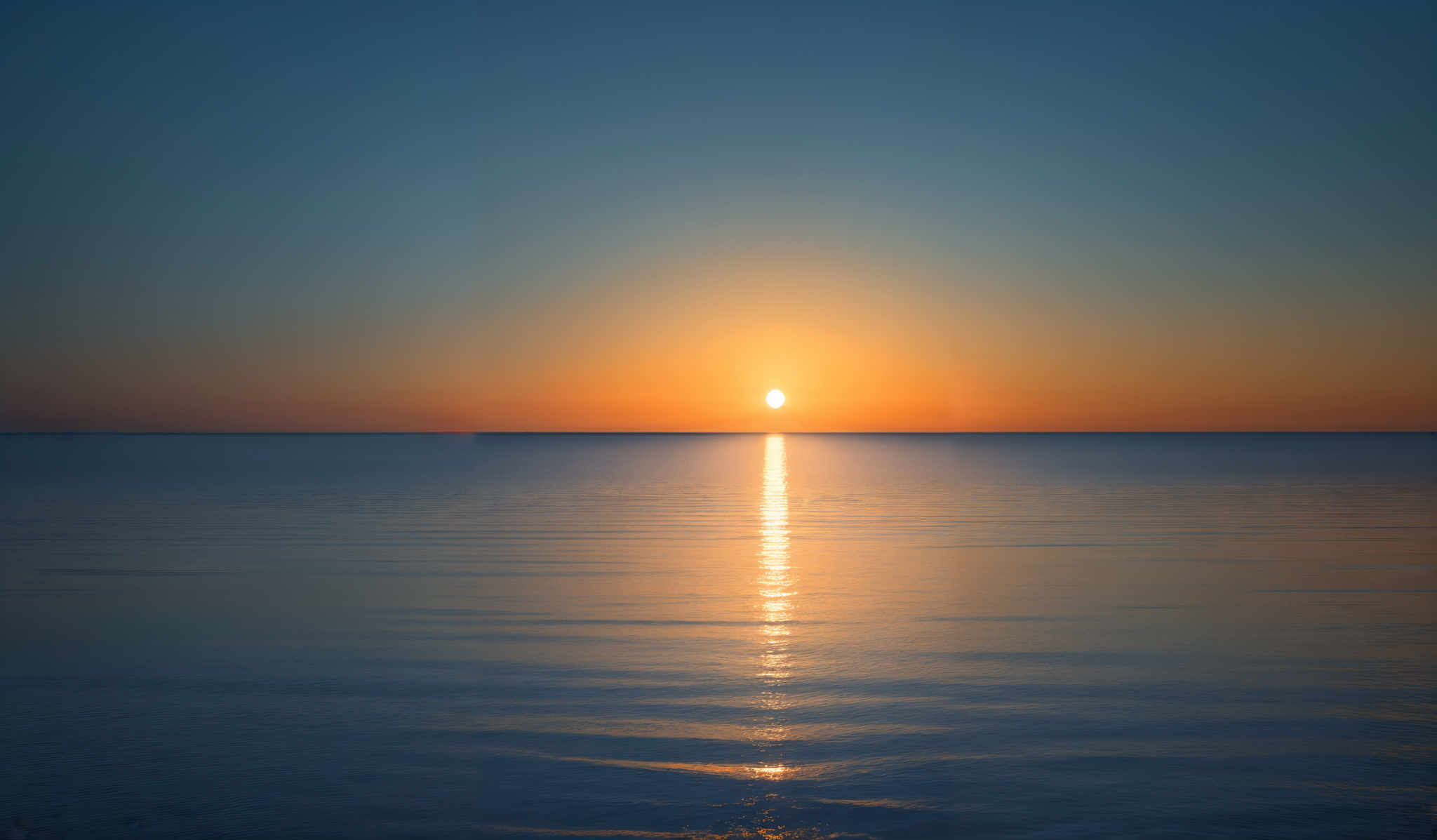 The image showcases a serene sunset over a calm body of water. The sky transitions from a deep blue at the top to a gradient of orange and yellow near the horizon. The sun is captured just above the waterline, casting a brilliant reflection that stretches across the water. Its golden hue contrasts beautifully with the darker shades of the water, creating a harmonious balance.