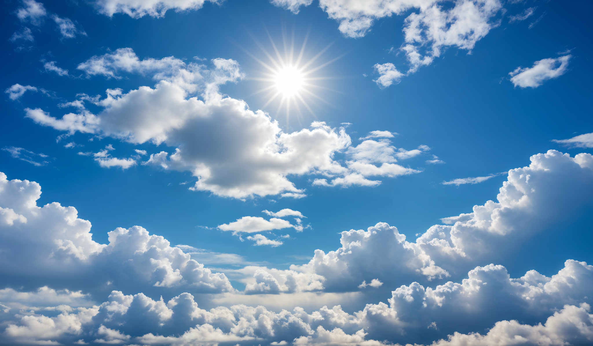 The image showcases a vibrant blue sky dotted with fluffy white clouds. The sun, radiating bright light, is positioned towards the top-center, casting a radiant glow. The clouds vary in shape, with some appearing as cumulus formations and others as more dispersed, wispy clouds.