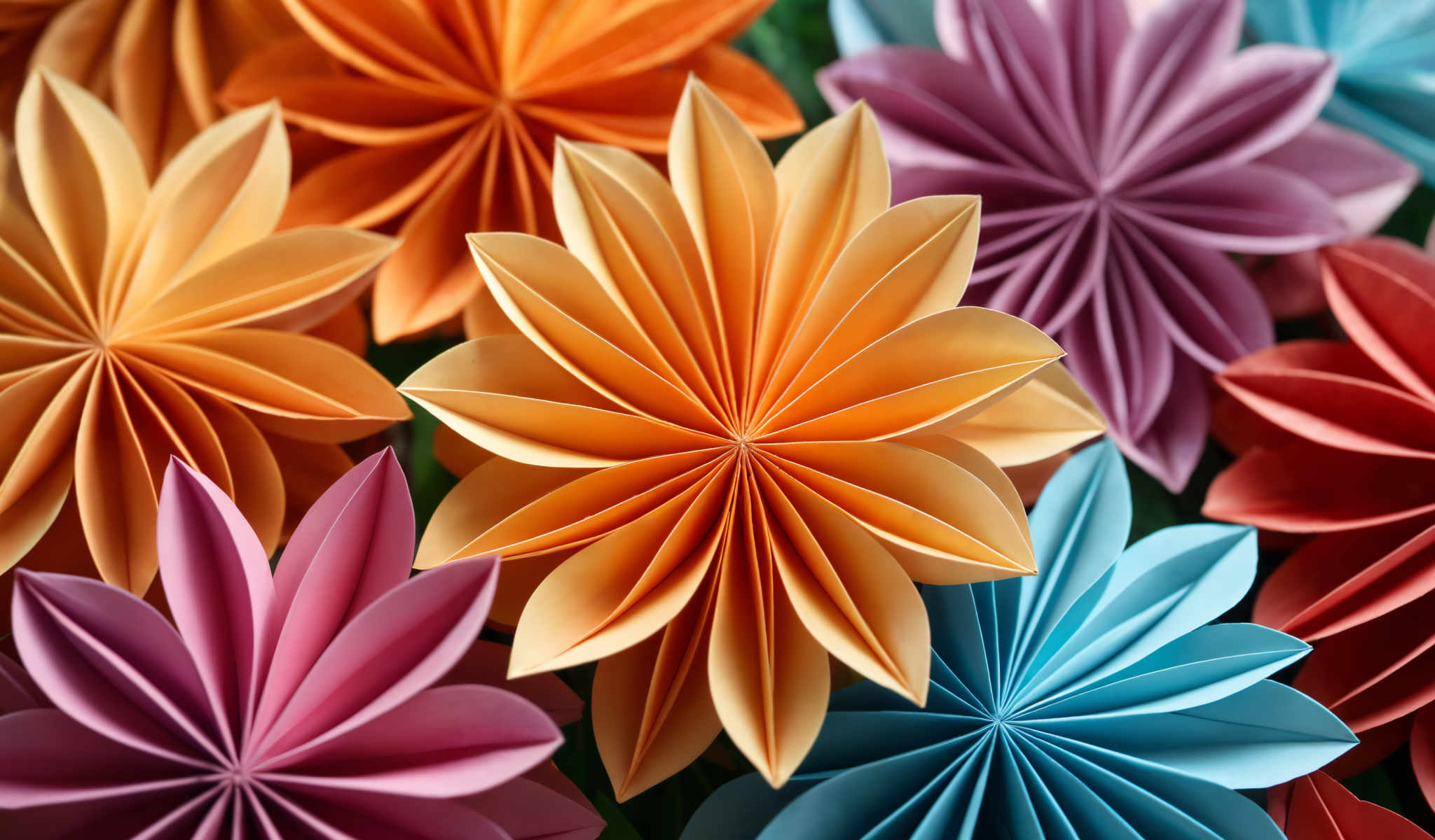 The image showcases a vibrant collection of origami flowers. The flowers crafted from paper are arranged in a circular pattern. Each flower is unique in color with hues ranging from orange and pink to blue and purple. The intricate folds and creases of the origami petals add a sense of depth and texture to the image making it a captivating visual experience. The overall composition of the flowers creates a harmonious balance of colors and shapes reflecting the beauty and precision of origamic art.