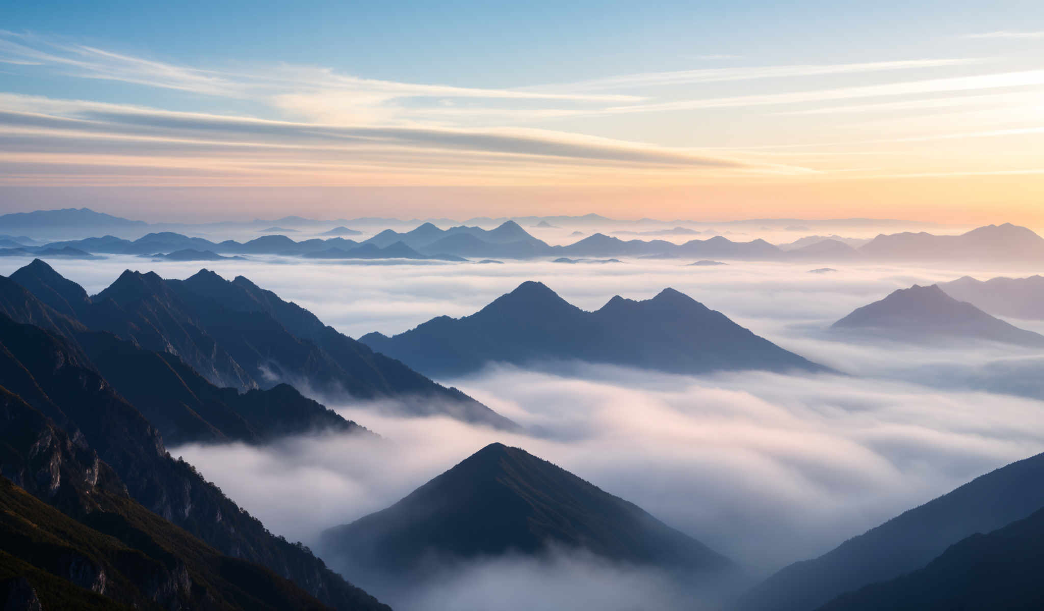 A breathtaking view of a mountain range with clouds in the foreground. The mountains are covered in a blanket of fog creating a serene and tranquil atmosphere. The sky above is a beautiful blend of orange and blue hues indicating that the photo was taken at sunrise. The perspective of the photo is from a high vantage point looking down on the mountains giving a sense of scale and grandeur to the scene. The image captures the majesty and beauty of nature in its purest form.