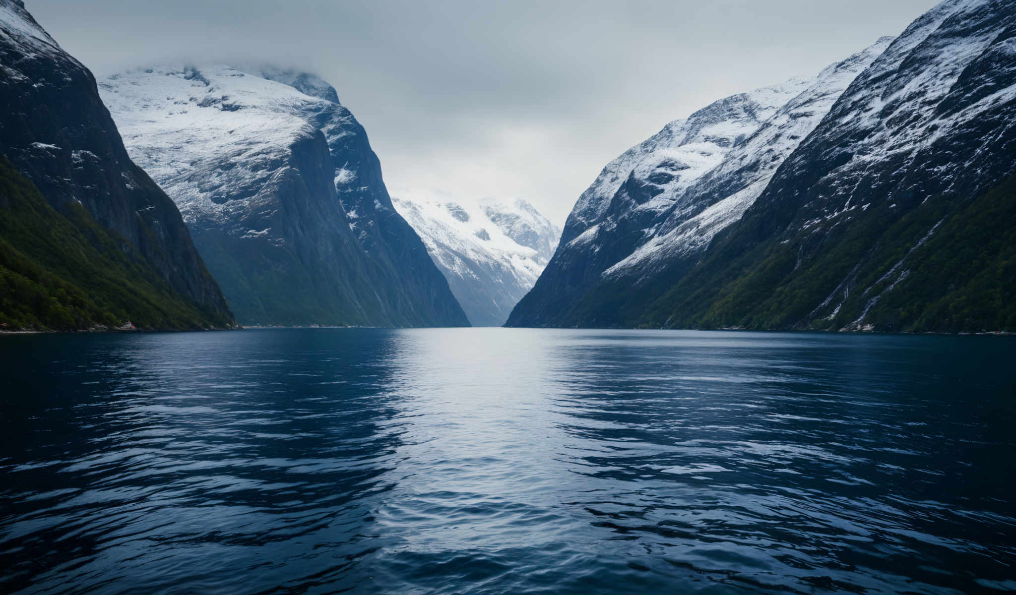 A serene scene of a mountainous landscape with a deep blue lake in the center. The mountains covered in snow rise majestically on either side of the lake their peaks obscured by a cloudy sky. The lake calm and still mirrors the surrounding scenery adding to the tranquility of the scene. The image captures the beauty of nature in its raw form untouched and unspoiled.