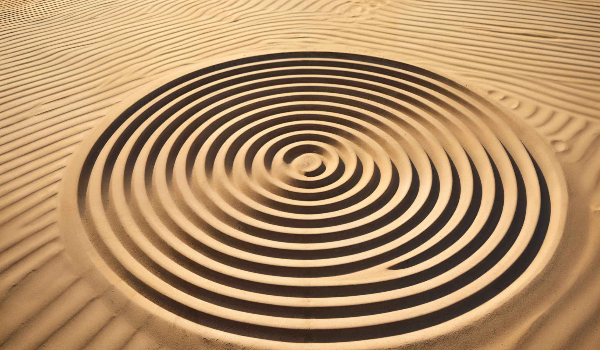 The image showcases a vast expanse of sand with intricate patterns. The dominant color is a warm beige, indicative of sand. The primary focus is on a series of concentric circles, each getting progressively tighter, creating a spiral effect. These circles are meticulously carved into the sand, with the sun casting shadows that accentuate the depth and dimension of the design. The ripples in the sand around the circles suggest the presence of wind or water, adding to the natural beauty and texture of the scene.