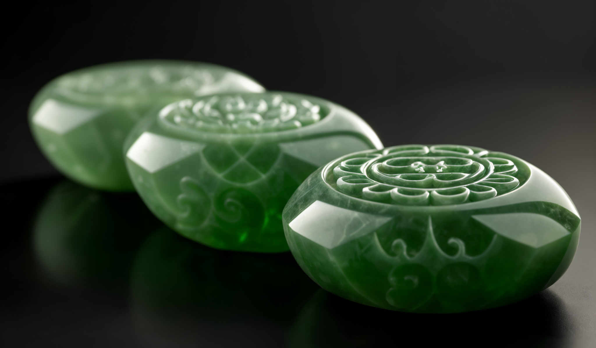 The image showcases three spherical objects with a translucent green hue. They are intricately carved with ornate patterns, predominantly featuring swirls and floral motifs. The carvings are symmetrical and appear to be deeply etched into the surface of the objects. The spheres are placed on a reflective surface, and the background is dark, emphasizing the objects' luminosity and detail.