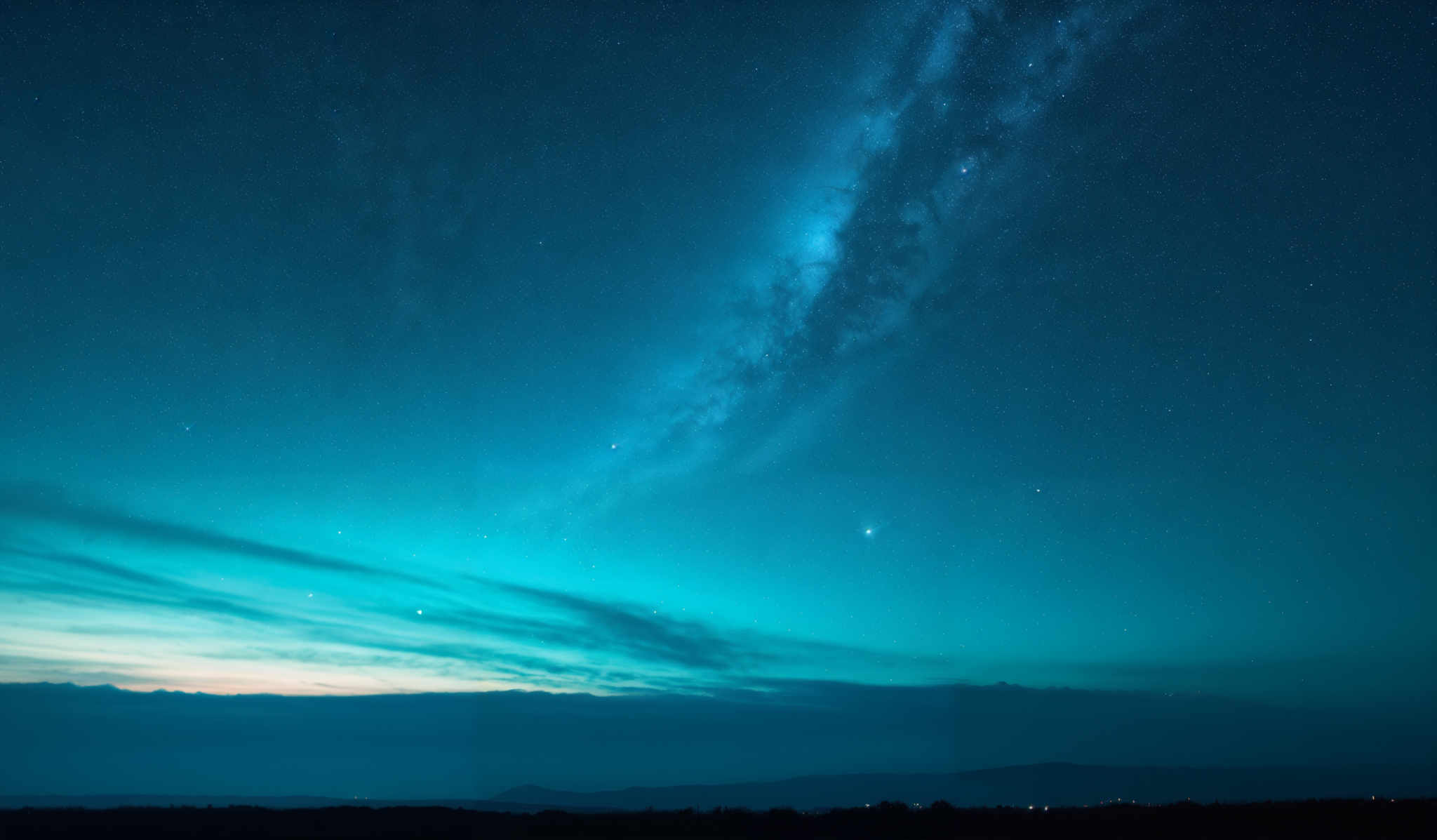 The image showcases a breathtaking night sky. The dominant color is a deep blue, transitioning to a lighter shade near the horizon. The Milky Way galaxy, with its dense cluster of stars, stretches across the sky, appearing as a milky band. The stars are scattered throughout, with some appearing as bright specks and others forming faint clusters. The horizon displays a gradient of colors, transitionning from a deep teal to a soft orange, possibly indicating the presence of distant city lights or the onset of dawn.