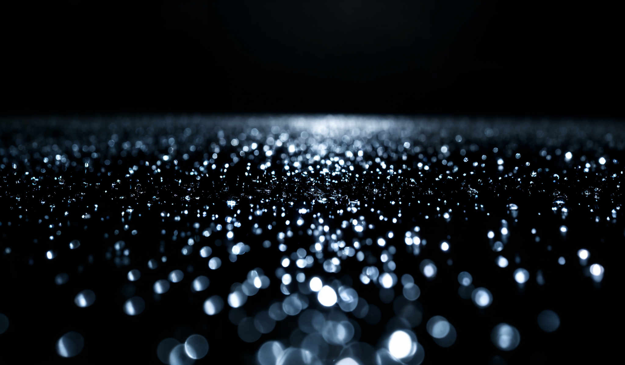 The image showcases a vast expanse of shimmering, glistening particles that appear to be suspended in the air. These particles reflect light, creating a mesmerizing pattern of light and dark spots. The predominant colors are deep blues and blacks, with the particles themselves emitting a bright, almost ethereal white light. The shape of the particles is varied, with some appearing as small dots and others as elongated specks, creating an effect reminiscent of a starry night or a galaxy.