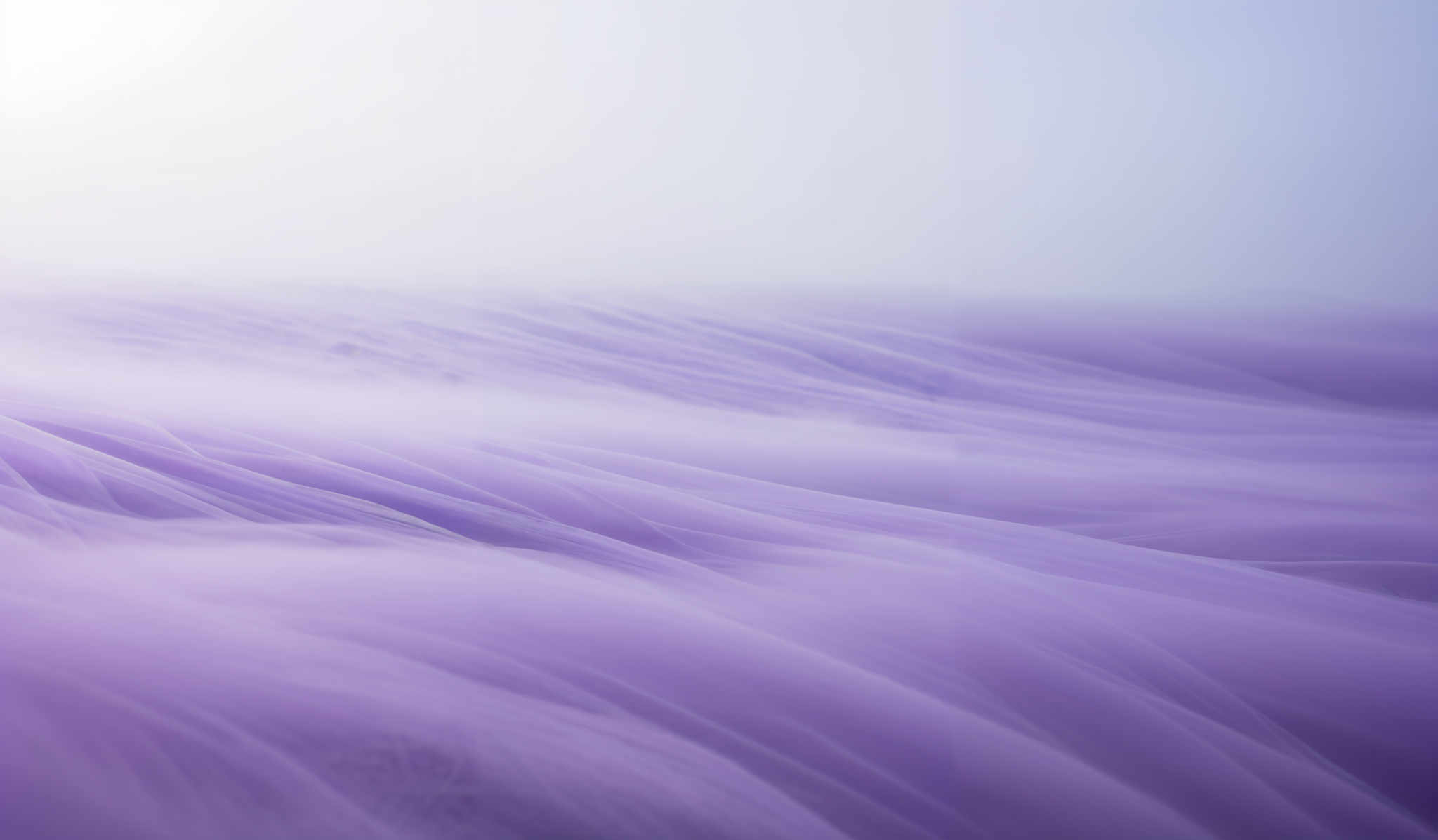 The image showcases a vast expanse of what appears to be a frozen landscape. The dominant color is a shade of purple, which gives the scene a dreamy and ethereal quality. The landscape is characterized by undulating patterns, reminiscent of flowing fabric or waves, which create a sense of movement and fluidity. The horizon is faintly visible, suggesting the vastness of the scene.