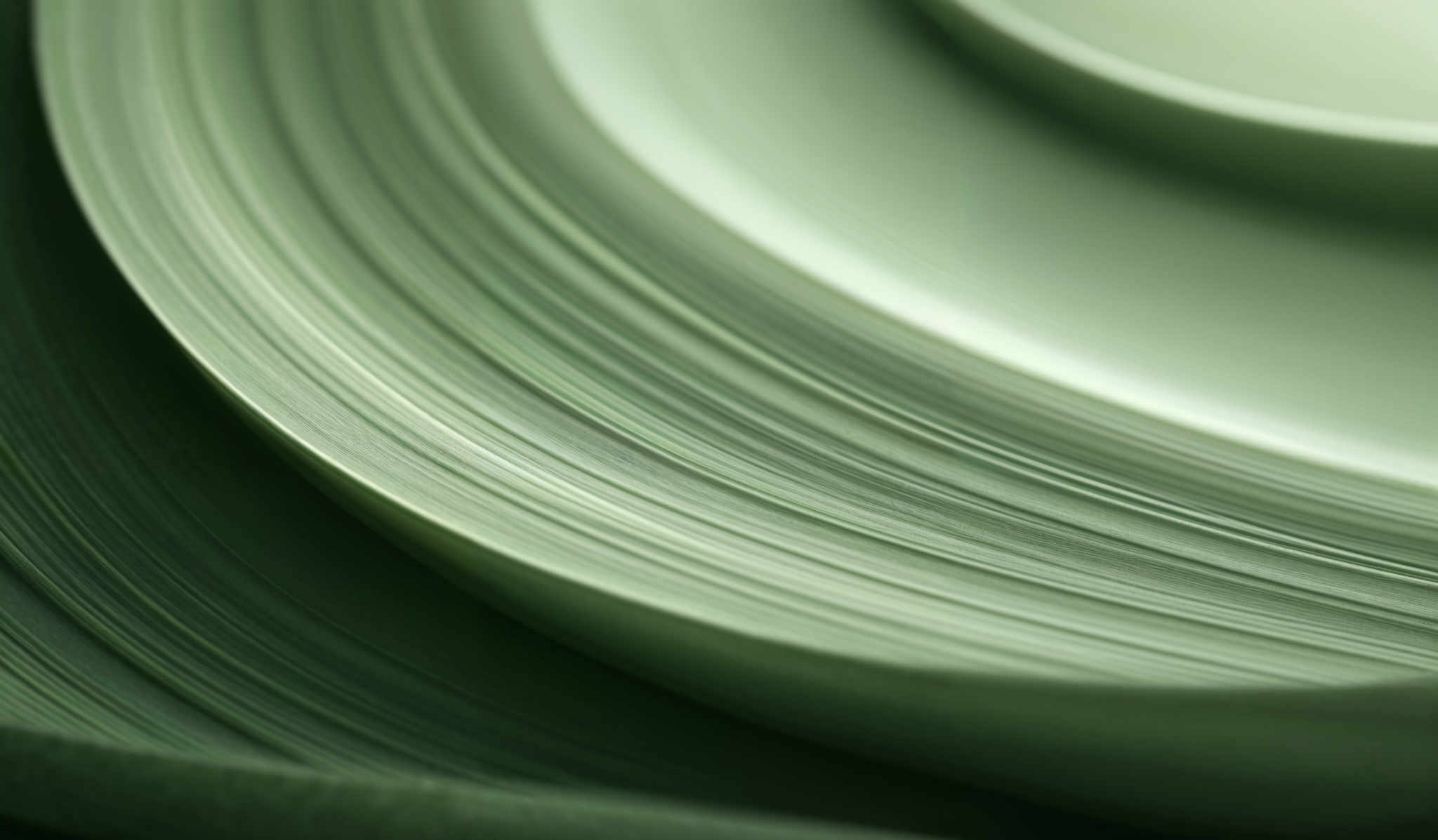 The image showcases a close-up view of concentric, curved lines that appear to be made of a material with a smooth texture. The dominant colors are varying shades of green, ranging from light to dark. The shapes are circular and semi-circular, with the lines appearing as if they are stacked or overlapping. The curvature of the lines gives a sense of depth and dimension to the image, making it appear as though the viewer is looking into a spiral or a ripple effect.