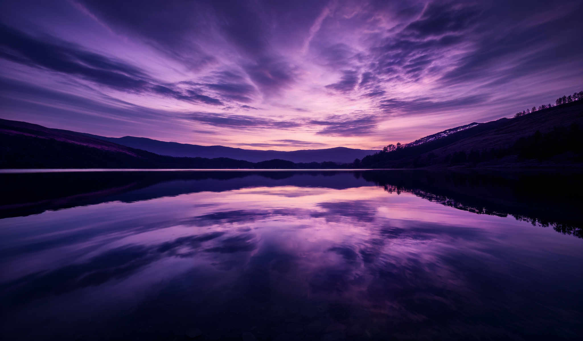 The image showcases a breathtaking landscape during what appears to be either dawn or dusk. The sky is painted with hues of purple, pink, and blue, with streaks of clouds that seem to be moving rapidly, creating a dynamic and dramatic effect. The reflection of the sky on the calm water below mirrors the colors and shapes perfectly, creating an almost symmetrical visual effect. In the foreground, there's a serene body of water that reflects the sky and the silhouette of the surrounding hills and trees. The hills are covered with trees, and their contours are visible against the backdrop of the vibrant sky.