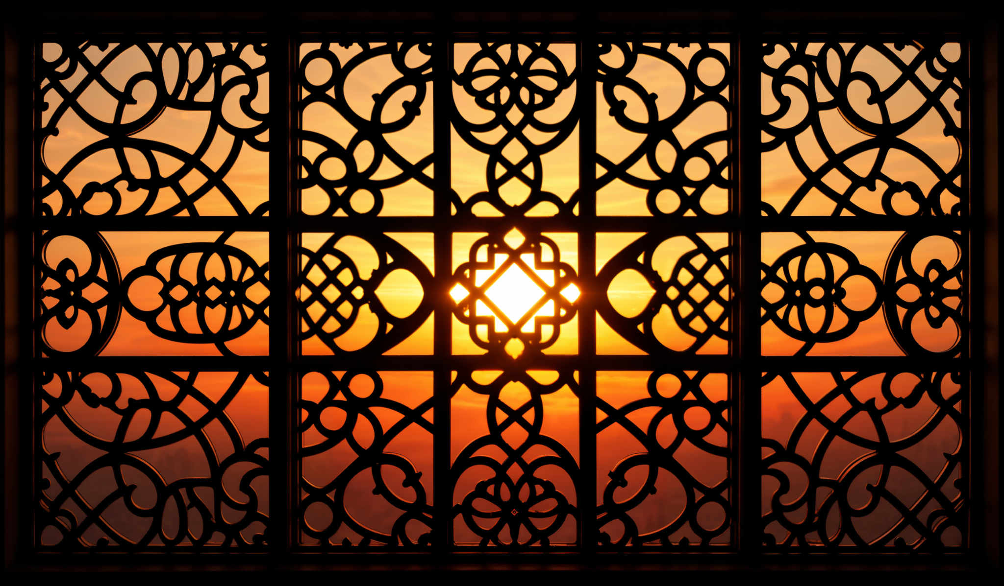 The image showcases a beautiful sunset or sunrise, with the sun casting a warm, golden hue. The sky is painted in shades of orange, pink, and purple. In the foreground, there's a window or door with intricate, ornate ironwork or metal grill patterns. These patterns are symmetrical and feature swirls, loops, and floral motifs. The sun is positioned in the center of the window, creating a radiant and captivating effect.