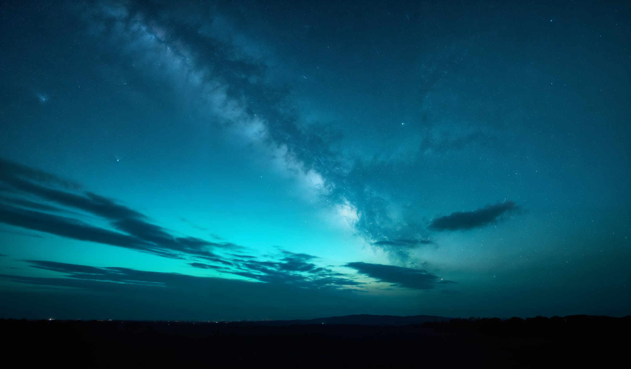 The image showcases a breathtaking night sky. The dominant color is a deep blue, transitioning to a lighter shade near the horizon. The Milky Way galaxy, with its dense cluster of stars, stretches across the sky, creating a milky band. There are also numerous stars scattered throughout the image, some of which are brighter than others. The horizon displays a mix of colors, with hints of green and turquoise, possibly indicating the presence of airglow or distant city lights. The sky is dotted with clouds, which appear wispy and ethereal, adding depth and dimension to the scene.