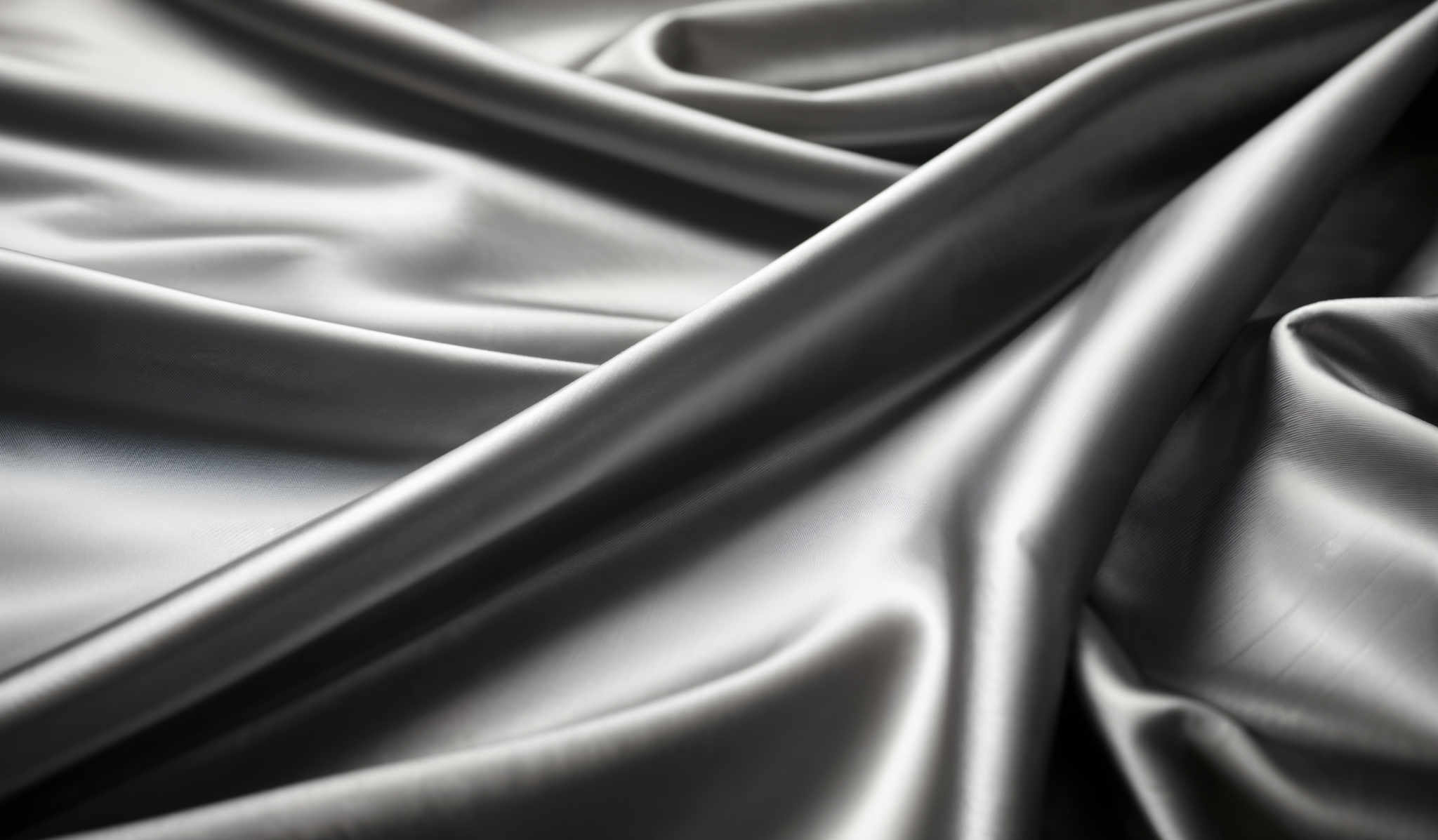 The image showcases a close-up view of a smooth, shiny fabric, possibly silk or satin. The fabric displays a gradient of colors ranging from a light gray to a deeper gray. The folds and creases in the fabric create a dynamic and flowing pattern, emphasizing its softness and luxurious feel.