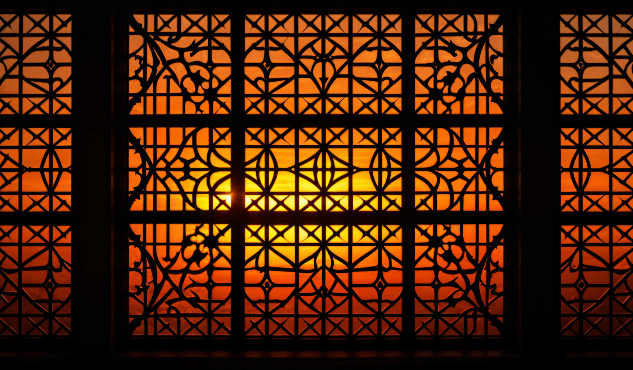 The image showcases a beautifully intricate window or grille design. The window is rectangular in shape, with a central square that is slightly larger than the surrounding rectangles. This central square is adorned with a detailed geometric pattern, consisting of interconnected triangles, diamonds, and other shapes. The entire design is set against a backdrop of a sunset or sunrise, with the sun casting a warm, golden hue that contrasts beautifully with the dark silhouette of the window. The colors range from deep blacks and dark browns of the metalwork to the bright oranges and yellows of the sky.