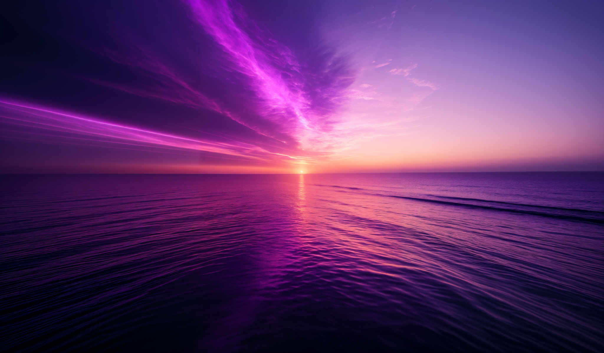 The image showcases a breathtaking sunset over a vast body of water. The sky is painted with a gradient of colors, transitioning from deep purples and blues at the top to warm oranges and yellows near the horizon. The sun is positioned just above the water, casting a brilliant reflection on the water's surface. The clouds are streaked with vibrant purple hues, adding to the ethereal beauty of the scene. The overall shape is expansive, with the sun setting in the distance and the water stretching out to meet the horizon, creating a sense of vastness and serenity.