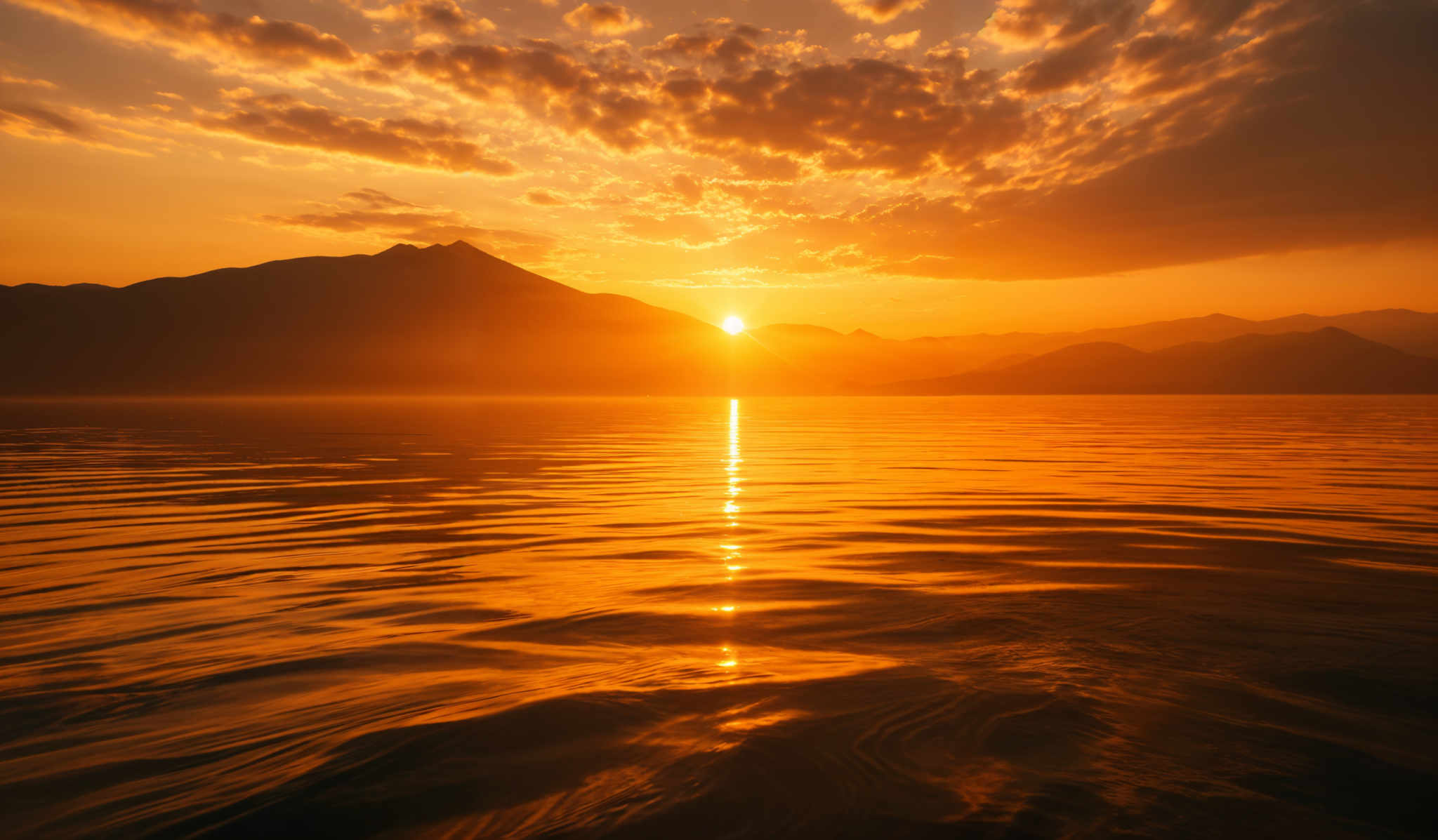 The image showcases a breathtaking sunset over a serene body of water. The sky is painted with hues of orange, gold, and deep red, interspersed with patches of dark clouds. The sun, positioned near the horizon, casts a brilliant golden reflection on the water's surface, creating a shimmering path of light. In the background, silhouetted against the fiery sky, are majestic mountain ranges. The water' s ripples add texture and depth to the scene, reflecting the colors of the sky and the mountains.