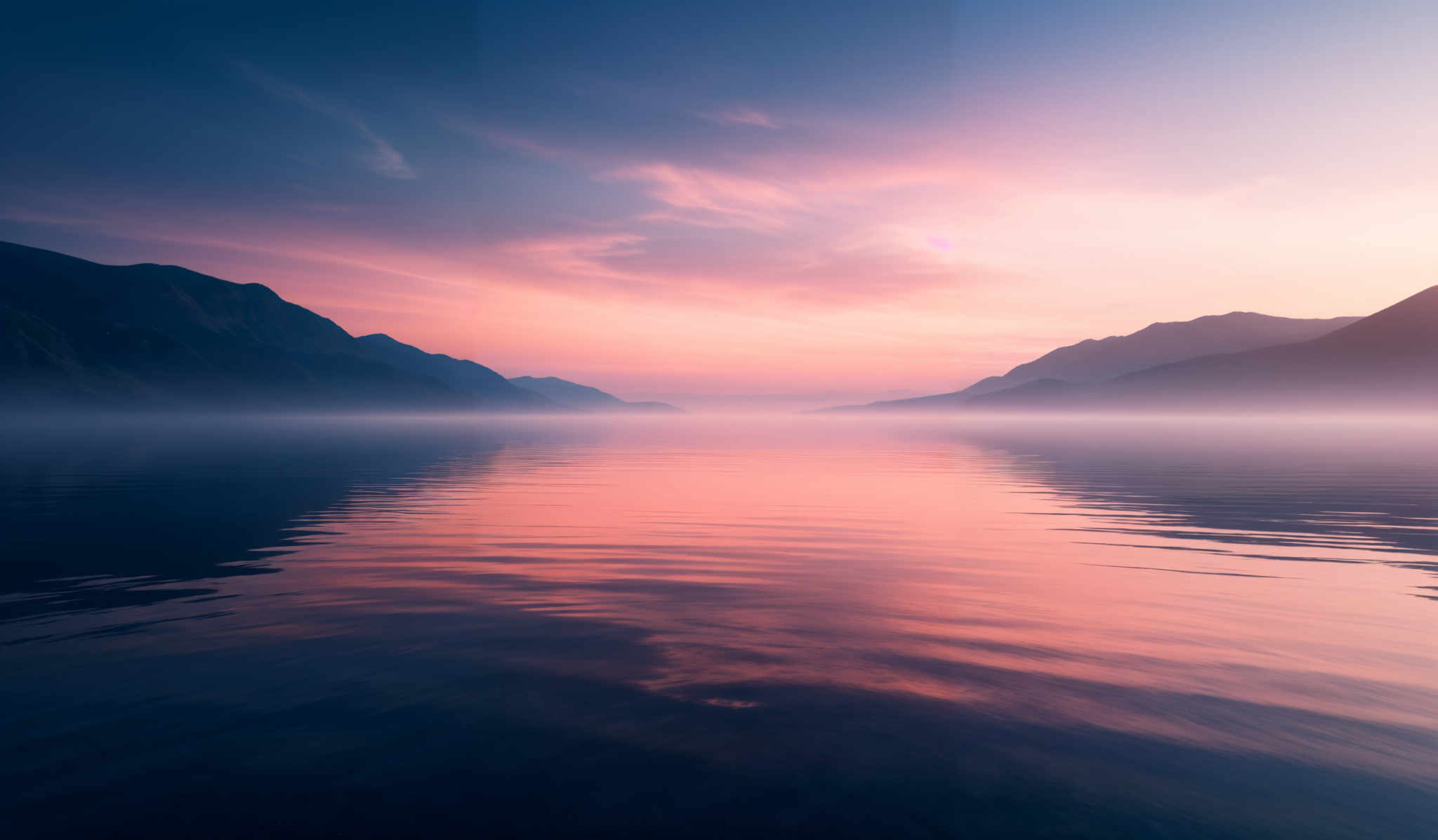 The image showcases a serene landscape during what appears to be either dawn or dusk. The sky is painted with hues of blue, pink, and purple, with wispy clouds scattered across. The mountains in the background are dark, silhouetted against the vibrant sky. The water in the foreground reflects the colors of the sky, creating a mirror-like effect. A thin layer of mist or fog settles over the water, adding to the tranquility of the scene.