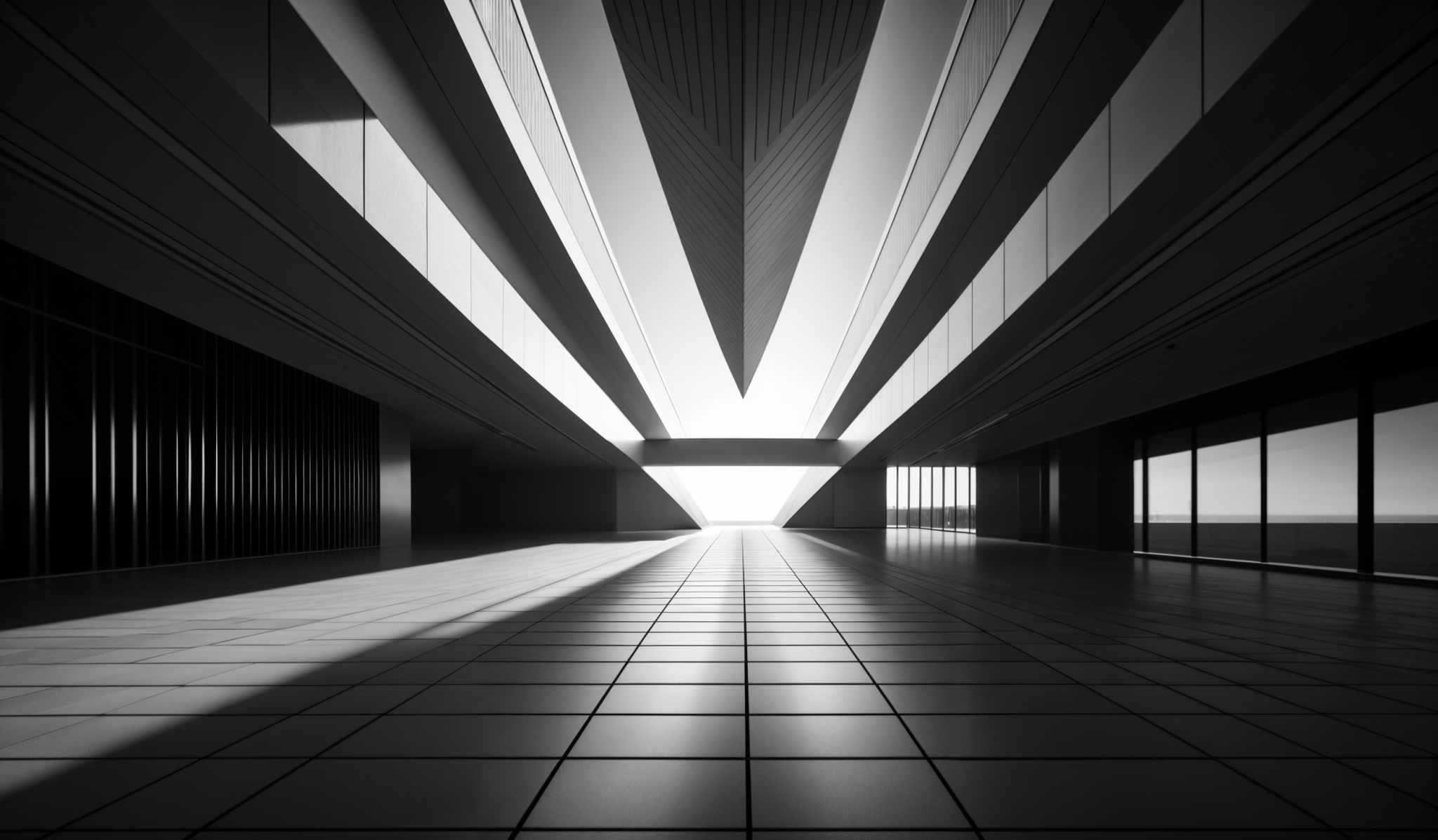 The image showcases a monochromatic, architectural space. Dominating the scene is a large, angular structure that appears to be a part of the building's design, with a sharp, triangular peak. This structure casts a pronounced shadow on the floor, creating a contrast with the illuminated areas. The floor itself is tiled, reflecting the light from the structure and the windows on the right. The walls have vertical lines, possibly indicating a modern design aesthetic. On the right side, there are large windows that allow natural light to flood in, further enhancing the play of shadows and light in the space.