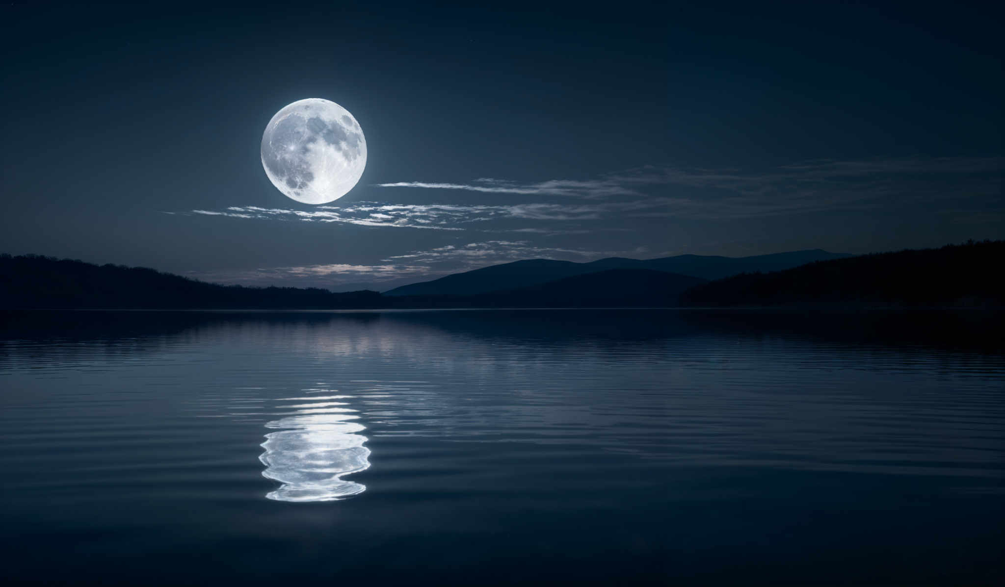 The image showcases a serene nighttime landscape. Dominating the scene is a large, luminous moon, which appears almost full, casting a bright glow over the scene. The moon's reflection is beautifully captured on the calm waters below, creating a mirror-like effect. The sky is painted in deep shades of blue, with wisps of clouds scattered around. In the distance, silhouettes of mountains can be seen, and the horizon is gently illuminated, possibly by the moon' or another light source. The overall color palette is cool, with the moon being the primary light source, casting shadows and highlights across the landscape.