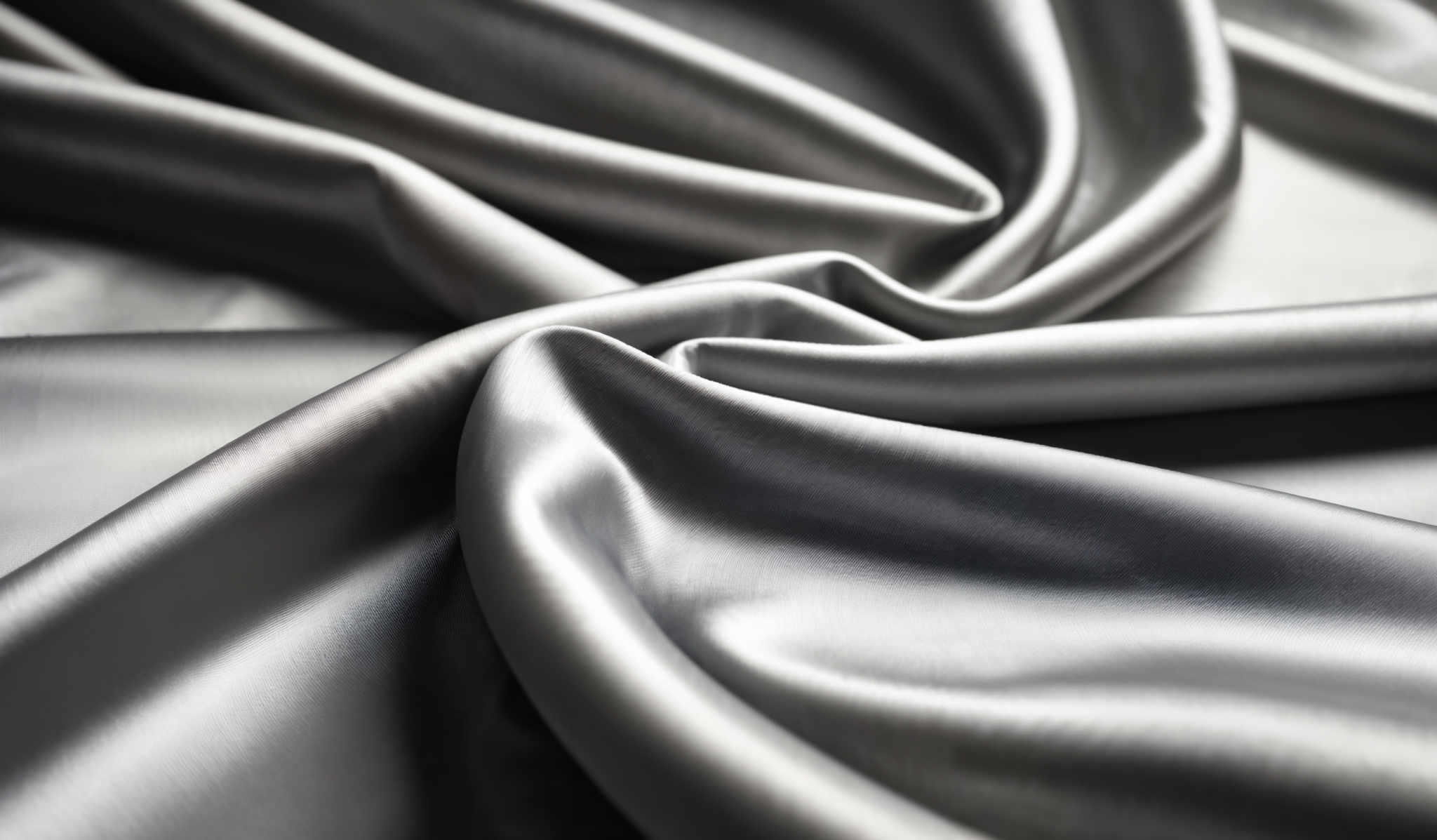 The image showcases a close-up view of a metallic or silky fabric with a shiny, reflective surface. The predominant color is a shade of silver or gray. The fabric displays smooth, wavy folds that create a pleasing contrast. The intricate details of the folds and the sheen of the material are captured beautifully, giving a sense of luxury and elegance.