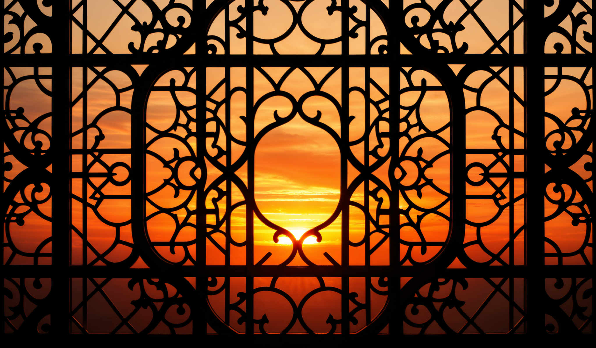 The image showcases a beautiful sunset with hues of orange, yellow, and pink. The sun is seen setting behind a series of intricate, ornate ironwork patterns. These patterns are symmetrical and feature swirls, loops, and floral motifs. The ironwork is set against a backdrop of a clear sky, and the sun's rays pierce through the gaps, creating a dramatic and aesthetically pleasing effect.