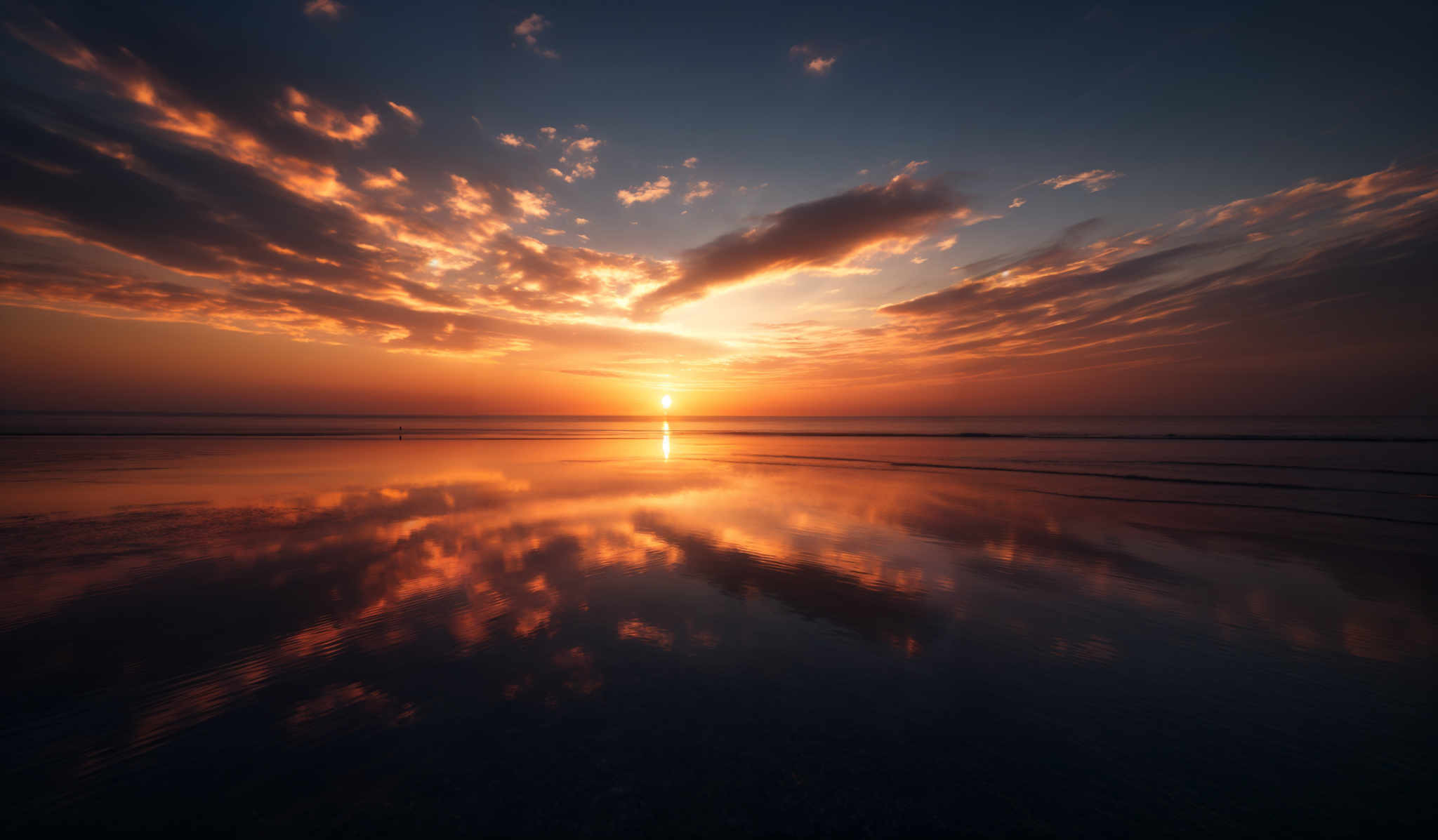 The image showcases a breathtaking sunset over a calm beach. The sky is painted with hues of orange, gold, and deep blue, with scattered clouds reflecting the sun's golden light. The sun itself is a brilliant orb, casting a brilliant reflection on the water below. The beach appears wet, possibly from a recent tide, and the water mirrors the sky's colors, creating a symmetrical view. In the distance, the horizon meets the vast expanse of the ocean.