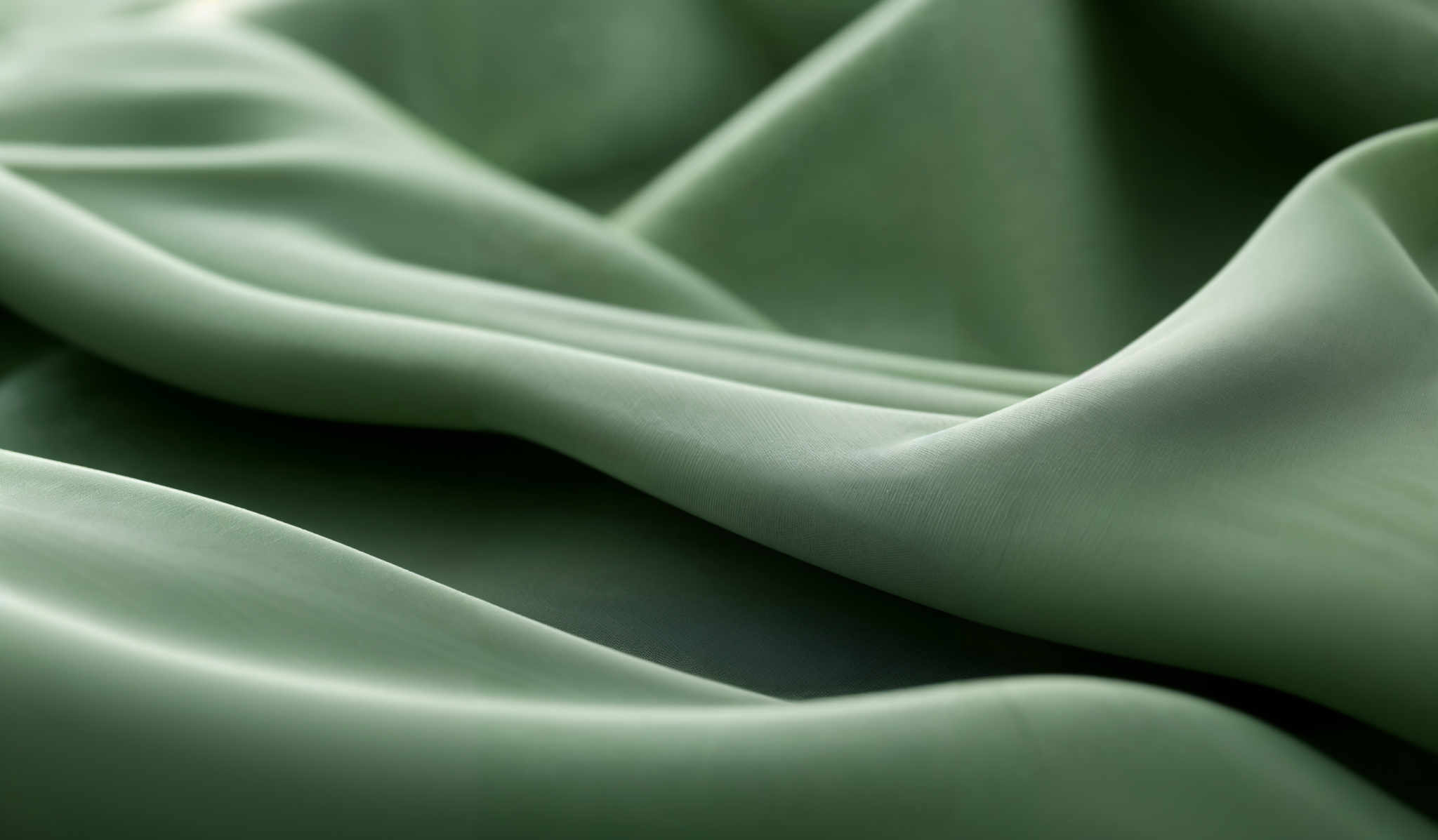 The image showcases a close-up view of a soft, flowing fabric in a shade of light green. The fabric displays gentle folds and creases, creating a wavy and undulating pattern. The texture appears smooth, with subtle variations in the sheen of the material, suggesting a delicate and luxurious fabric, possibly silk or satin.