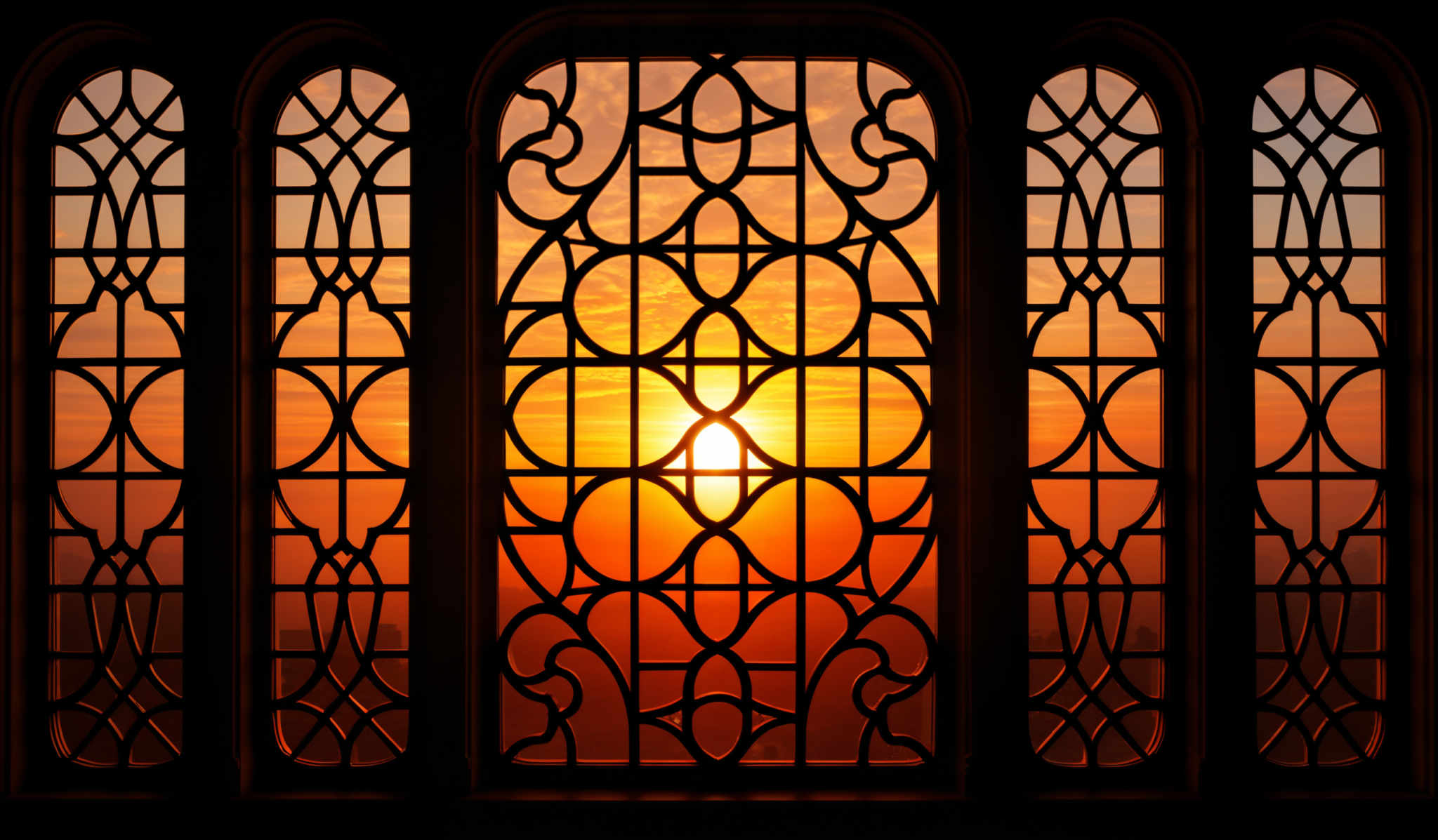 The image showcases a beautiful sunset viewed through an intricately designed window. The window features a symmetrical, ornate pattern with interconnected circles and curves. The sun, positioned centrally, casts a warm, golden hue that illuminates the sky with shades of orange, pink, and purple. The silhouette of the window's design is contrasted against the vibrant backdrop of the sunset, creating a visually stunning effect.