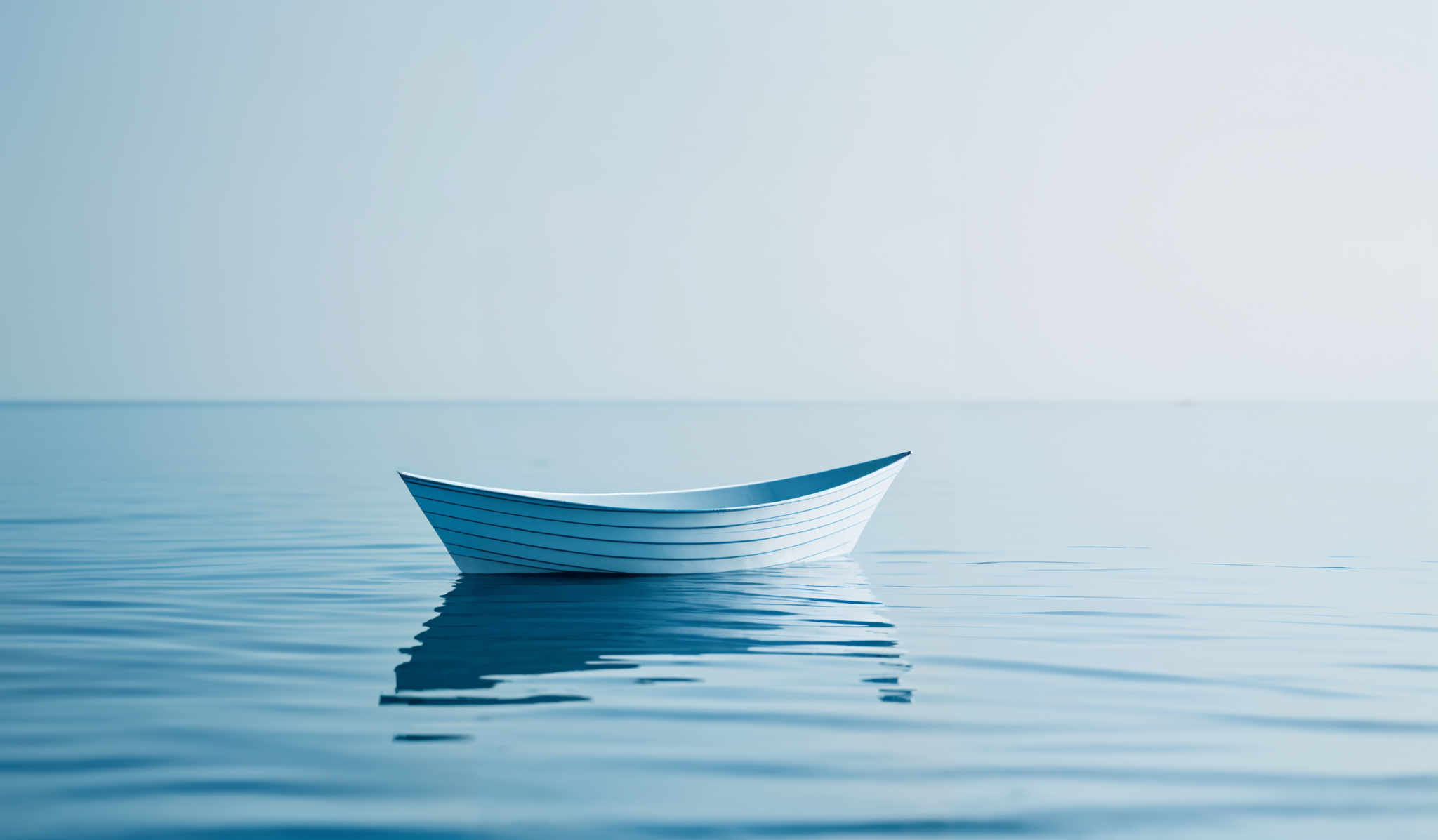 The image showcases a serene scene of a calm body of water, possibly an ocean or a large lake. Dominating the foreground is a solitary wooden boat, painted in a light blue hue. The boat's reflection is visible on the water's surface, adding depth to the image and emphasizing the tranquility of the scene. The water itself is a deep shade of blue, with gentle ripples that suggest a gentle breeze or the movement of small aquatic life. The horizon is faintly visible, merging the water with the sky, which is a soft, pale blue, adding to the overall peaceful ambiance of the image, which evokes feelings of solitude and contemplation.